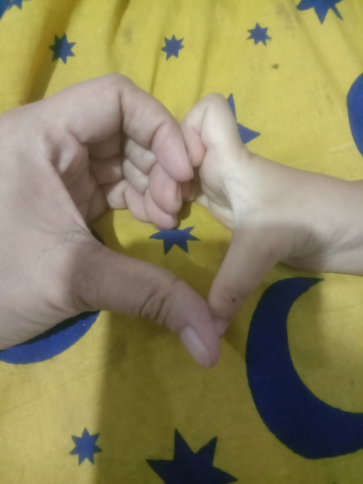 The image shows two hands, presumably from different individuals, forming a heart shape against a yellow fabric background with blue star and crescent moon patterns. The perspective is a close-up shot, with the hands occupying the majority of the frame. The fabric appears to be a patterned textile, possibly a blanket or clothing item. The lighting suggests an indoor setting. No specific landmarks or urban features are visible to determine its location within Cianjur, Indonesia. The hands are positioned centrally, with the thumbs touching and the index fingers and middle fingers forming the curves of the heart. The skin tones suggest a contrast between the two individuals. The overall scene is simple and focused on the gesture of forming a heart with the hands.