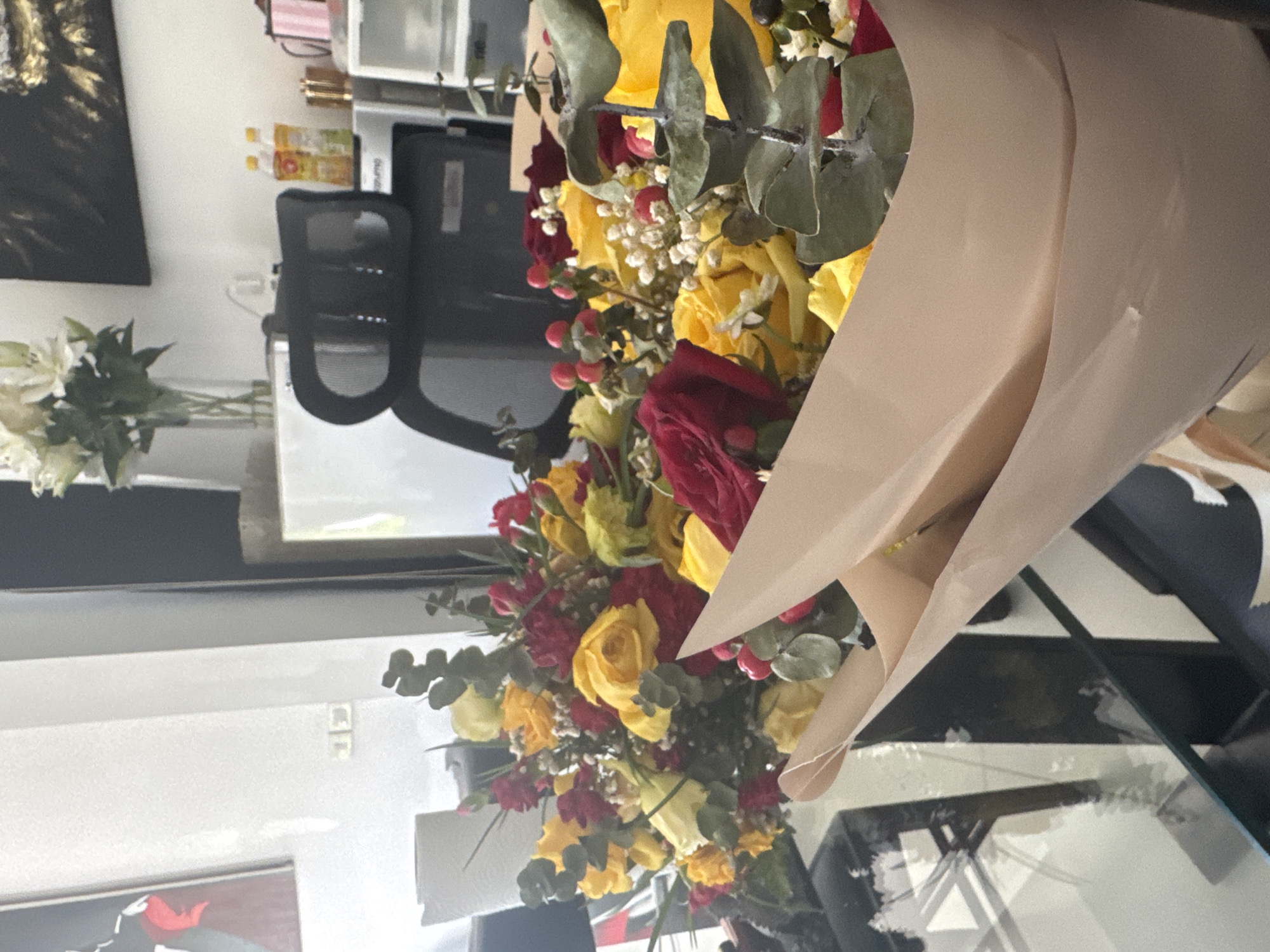 The image shows a close-up of a bouquet of yellow and red roses, along with some greenery and small red berries, resting on a glass table. The flowers are partially wrapped in tan paper. In the blurred background, a desk with a black office chair, bottles of drinks, and what appears to be a printer or scanner are visible. The overall scene suggests an indoor setting, possibly an office or home environment, located in Ko Samui District, Thailand. There are no people visible in the image.