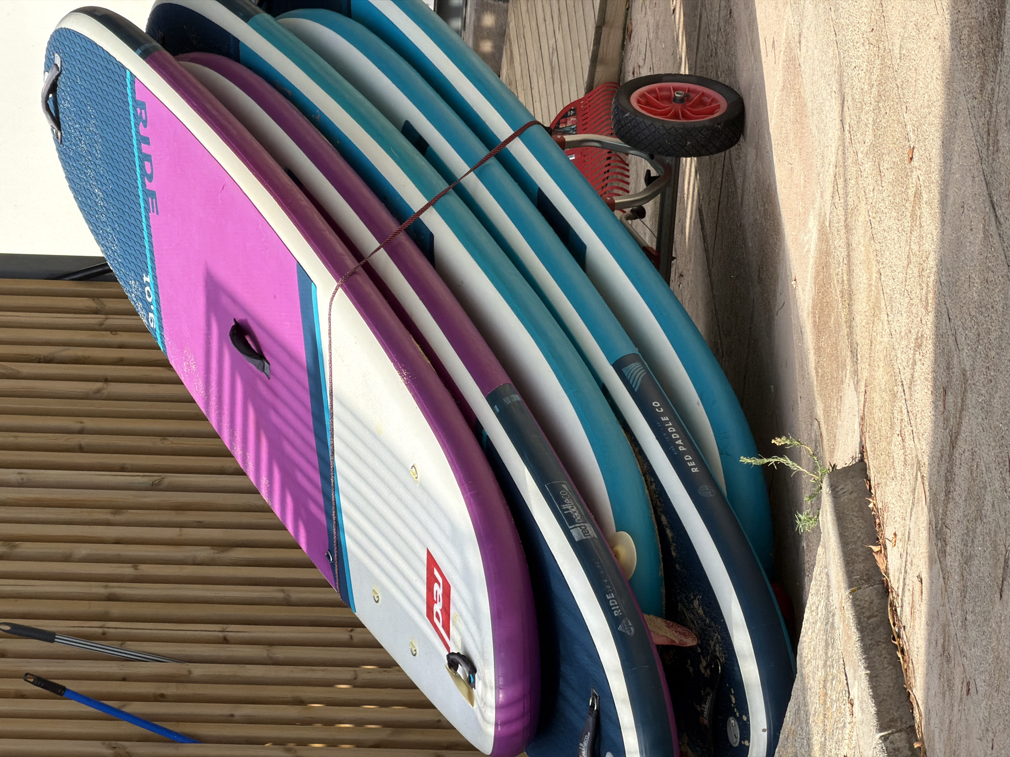 A stack of five inflatable stand-up paddleboards (SUPs) rests on a red wheeled cart, positioned outdoors near a wooden fence. The SUPs vary in color, with the top board being predominantly blue and pink, the second blue and white, the third blue and purple, and the bottom two boards being primarily blue and white. A thin red rope is wrapped around the stack of boards to secure them. The cart has two red wheels. To the left of the SUPs, leaning against the fence, are two cleaning tools: a blue-handled broom and a black and silver-striped brush. The ground is paved with concrete. A small patch of green vegetation is growing from the ground near the curb in the foreground. The scene is illuminated by sunlight, casting shadows from the fence and the SUPs onto the ground. The location is identified as Magaluf, Spain.
