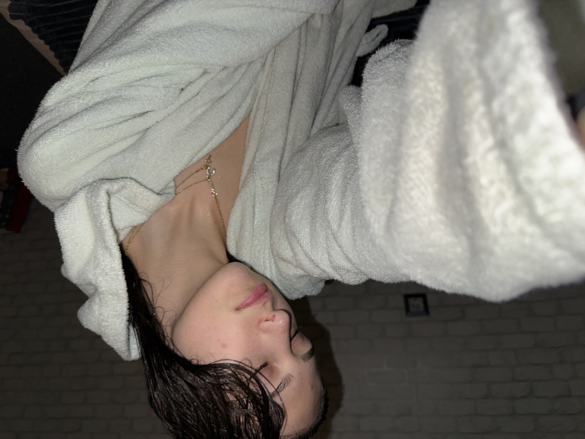 A close-up, upside-down view depicts a person with wet, dark hair and closed eyes, wearing a white terrycloth robe and a delicate gold necklace. The background appears to be a dimly lit brick wall. There's no indication of this image being in Sofia, Bulgaria.