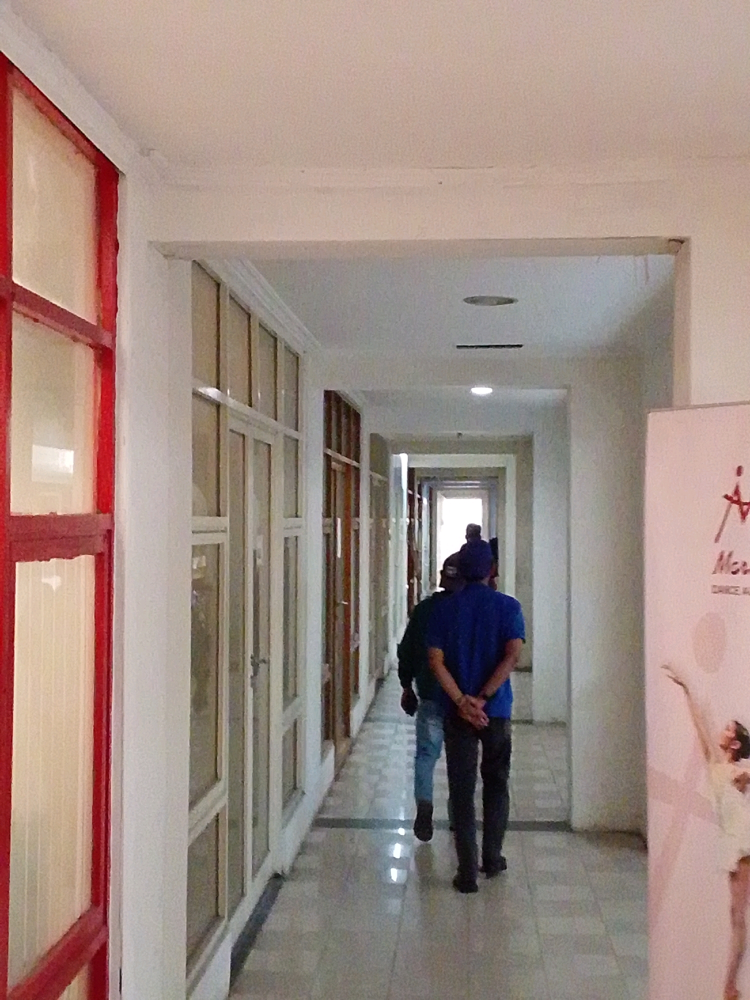 In a brightly lit hallway, two individuals walk away from the camera. The person in the foreground, wearing a blue t-shirt and dark pants, has their hands clasped behind their back. They are walking on a checkered tile floor. To their left is a wall with a large window framed in red. To their right, a large poster depicts a woman in a dancing pose. The hallway continues into the distance, with more doors and windows lining the walls. The environment appears to be indoors, possibly a school or an office building. The image does not provide specific clues to pinpoint a location within North Jakarta, Indonesia, but the overall setting suggests an urban or suburban indoor space.