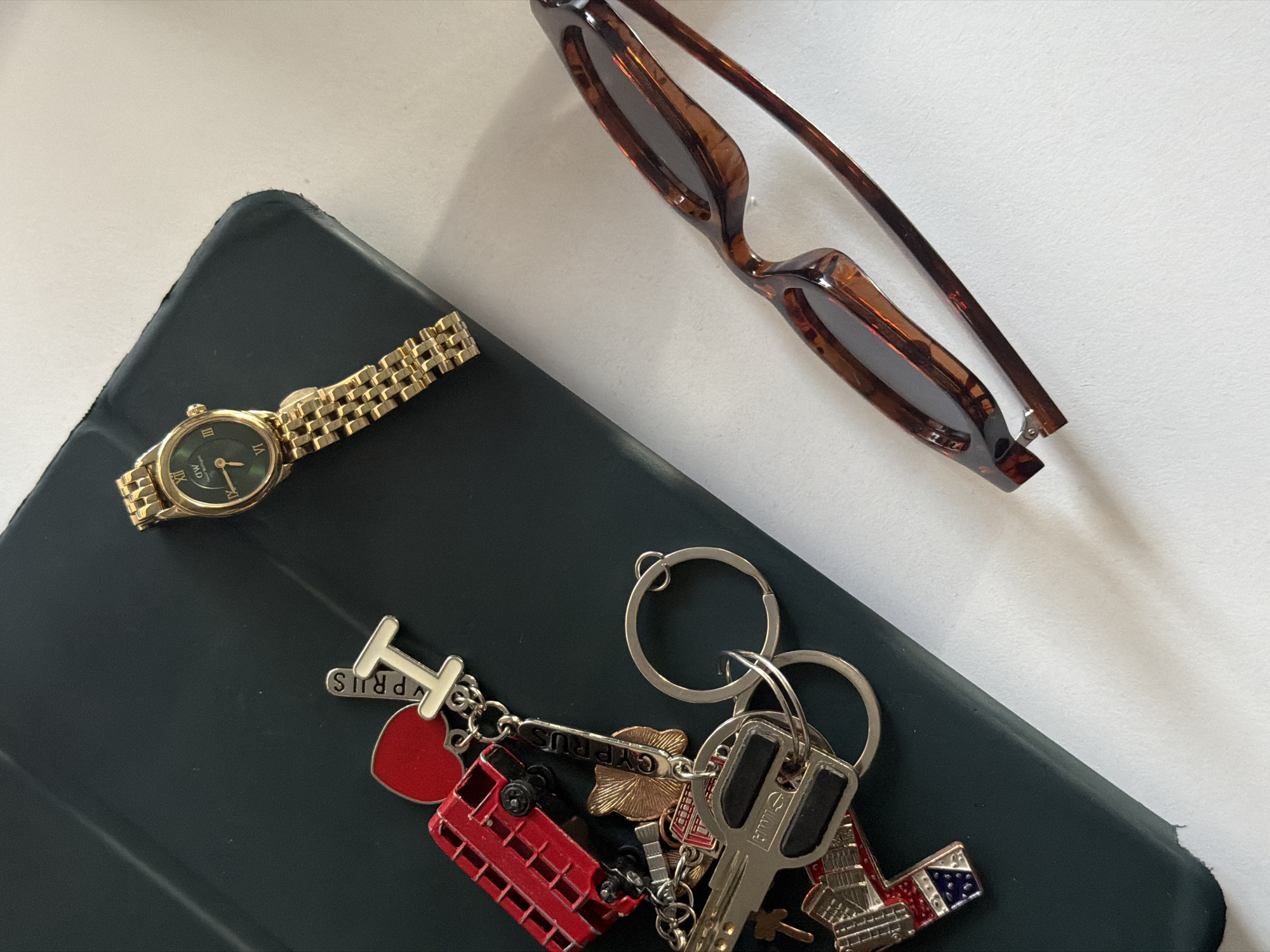 This overhead flat lay image captures a collection of personal items arranged on a dark, matte surface, likely a phone case or notebook, against a white background.
The objects include a gold-colored watch with a dark green oval face, featuring Roman numerals at the XII, III, VI, and IX positions, and the brand "MDL" visible on the dial. Adjacent to it are a pair of folded sunglasses with brown tortoise-shell frames.
A cluster of keys is attached to several silver keyrings. These keys are accompanied by a variety of souvenir keychains. One prominent keychain spells out "I ❤️ CYPRUS," with the heart symbol rendered in red. Another is a miniature red double-decker bus, with the word "LONDON" visible along its base. A gold-colored, sun-shaped charm also bears the text "CYPRUS" (inverted in the image). Other keychains include a miniature Statue of Liberty and another depicting a building or monument with a red, white, and blue pattern. One of the metal keys has partially visible text that appears to be "CILL".
The arrangement presents a still life of travel mementos. While the image was taken in Yerevan, Armenia, the keychains clearly point to past visits to locations such as Cyprus and London, and potentially other international destinations. No people are visible, and no specific activity is taking place. The time of day and weather conditions are not discernible from the image.