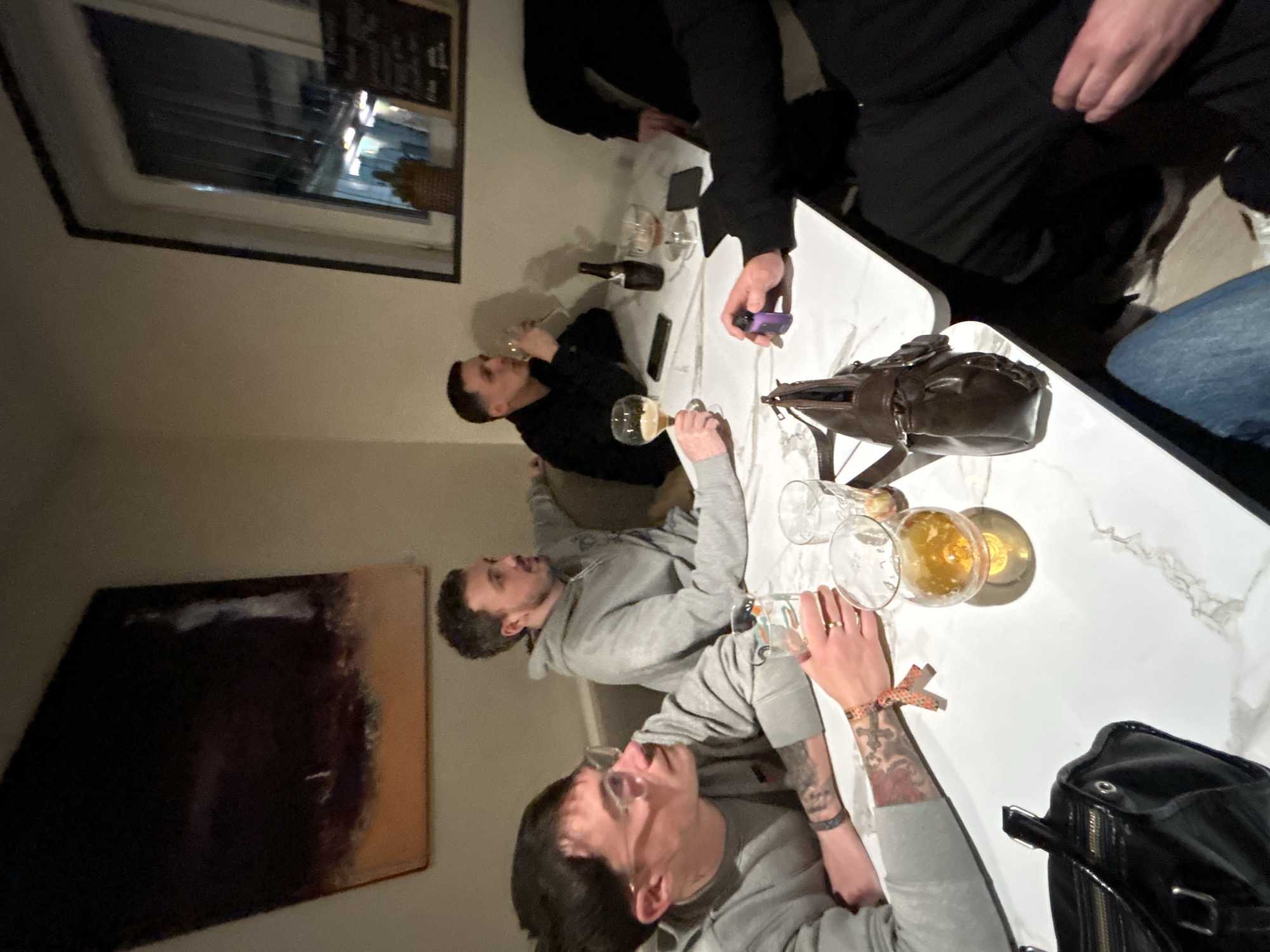 The image shows a group of people at a table. The table is white and has a marble-like pattern. There are several glasses on the table, some filled with a golden-colored liquid, likely beer. There's also a dark brown bottle and a dark-colored bag on the table. One person is wearing a black outfit, while others are wearing grey hoodies. One individual is shown drinking from a glass. Another individual has visible tattoos on their arm. The setting is indoors, and a window is visible in the background. The image provides no information about the location being in Hem, France.