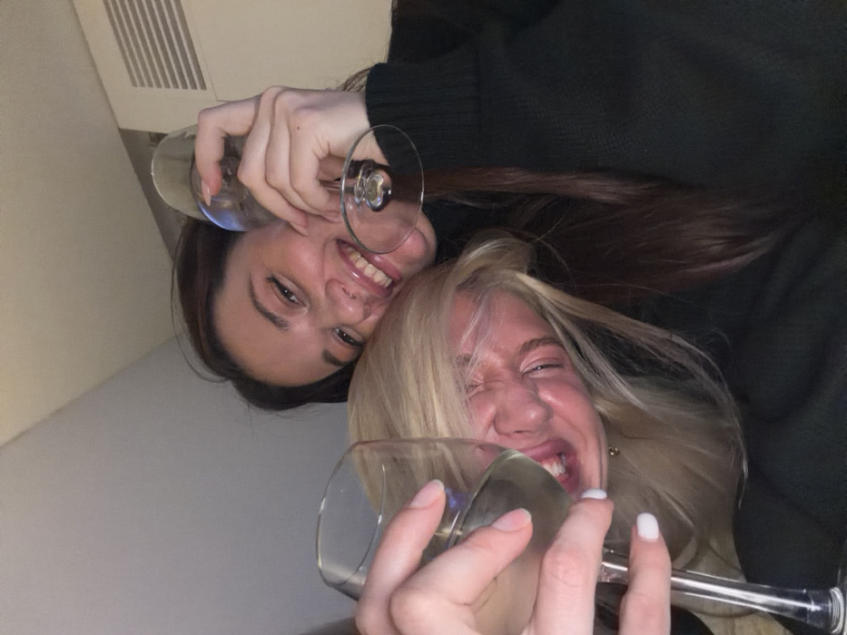 Two women hold wine glasses upside down, smiling. One woman has dark brown hair and a bright, open smile, while the other has blonde hair, squinted eyes, and a more playful grimace. Both are casually dressed in dark tops. The background includes a plain wall and what appears to be a radiator. The image has a casual, selfie-like feel, captured at a tilted angle.
