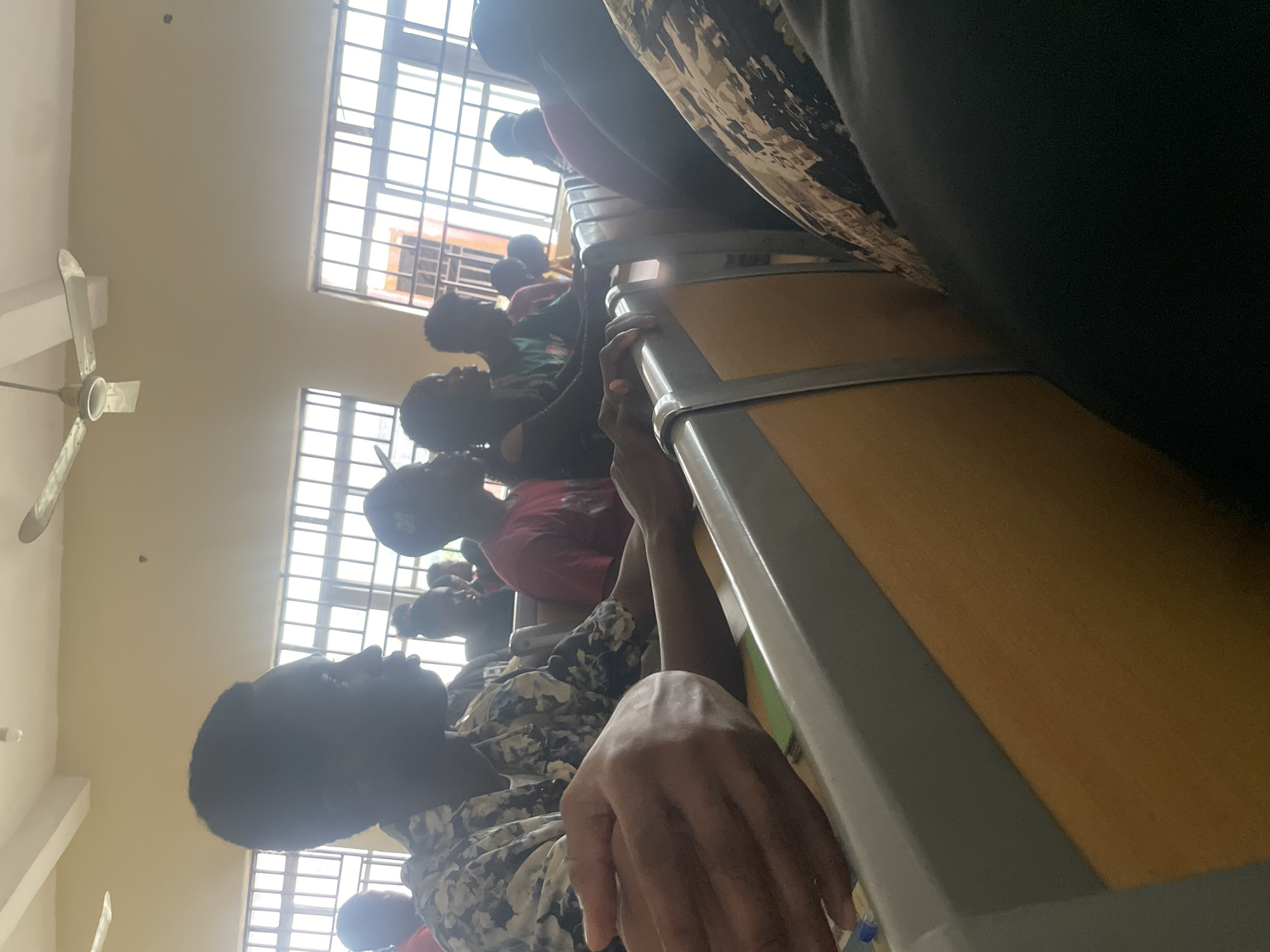 The image shows a group of young people, likely students, seated in a classroom or lecture hall. The setting appears to be in Ihiagwa, Nigeria, as suggested by the windows with barred panes typical of buildings in the region. In the foreground, a person wearing a camouflage shirt is visible from the side, with their hand resting on a desk. Other individuals are seated behind, some looking forward, suggesting they are engaged in a lesson or presentation. A ceiling fan is visible in the upper part of the image, indicating a warm climate. The overall scene depicts a typical educational environment.