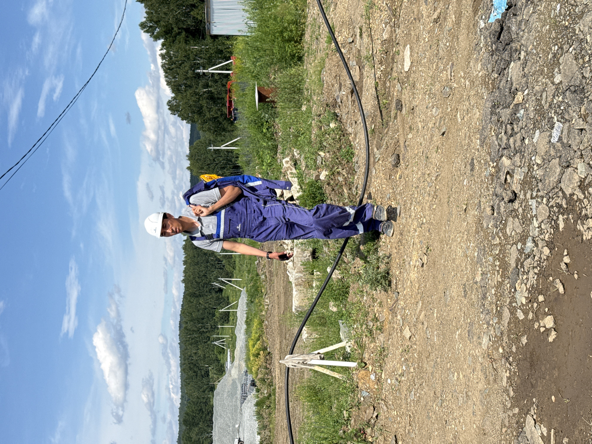 In Leninsk, Russia, on a sunny day, a man wearing blue overalls and a white hard hat walks on a dirt path in a construction area. He carries a blue backpack over his right shoulder and a folded blue and gray jacket over his left. The background features a line of trees, a large pile of gray gravel, and a white corrugated metal structure. A thick black cable runs across the foreground. The ground is a mix of dirt, gravel, and some puddles. The sky is blue with scattered white clouds.