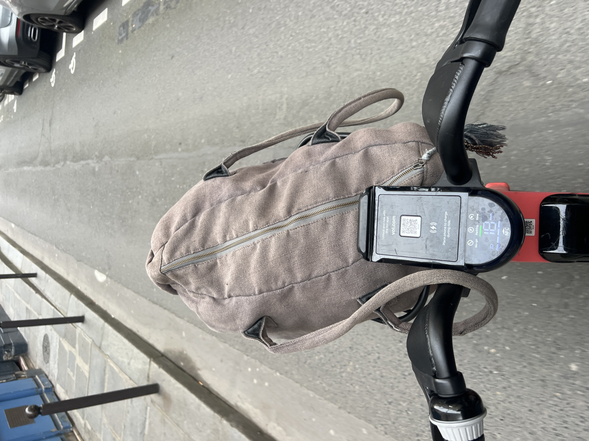 The image shows a top-down view of the handlebars of an electric scooter with a large canvas bag resting on them. The handlebars are black and have a digital display showing a speed of 1.9 km/h and a QR code for unlocking the scooter with the Voi app. The bag is a muted brown or gray color and has two handles. The scooter is on a street in Paris, France, as evidenced by the Parisian-style sidewalk and street. Cars are parked on the left side of the street.