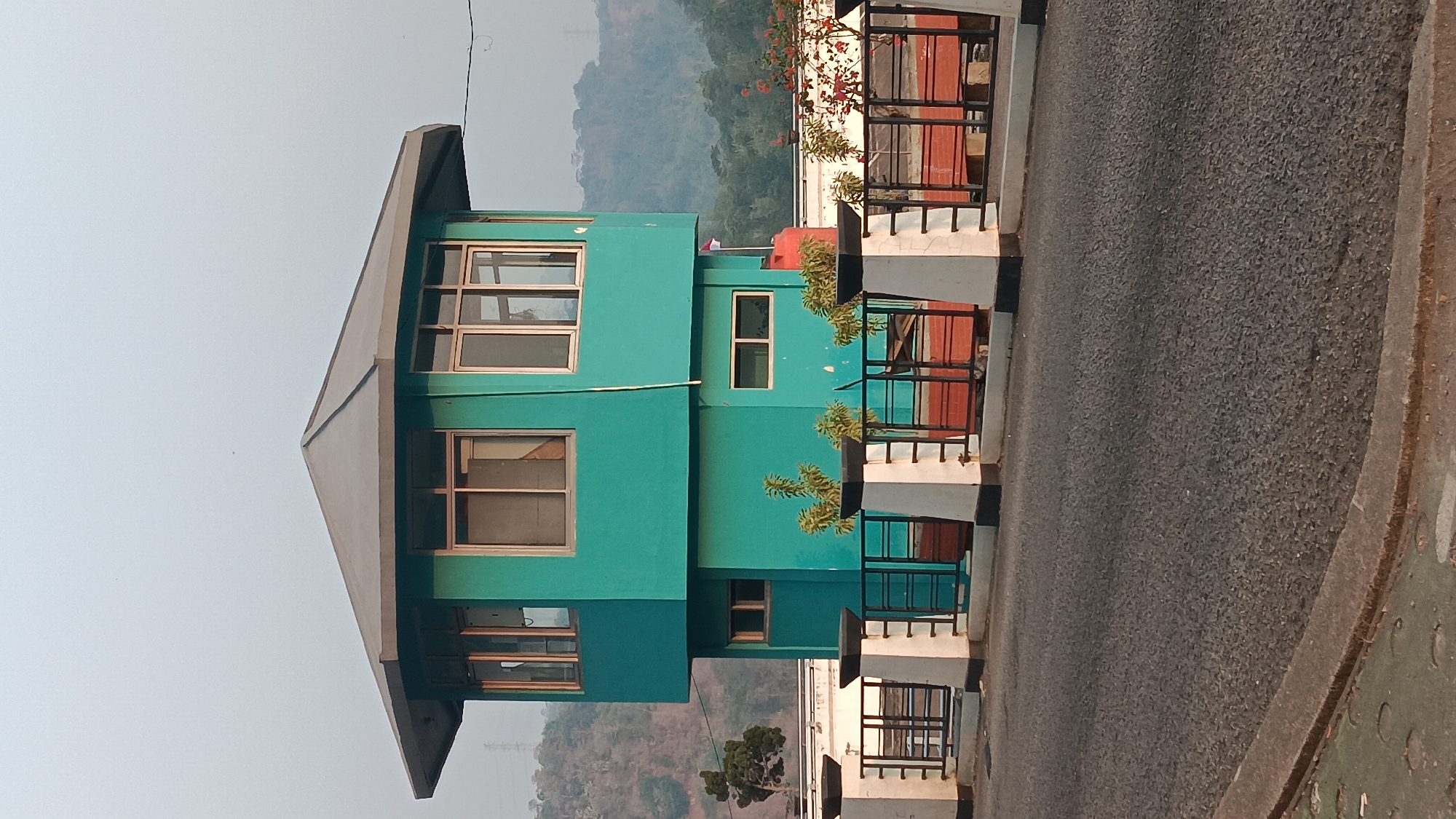 The image shows a hexagonal, turquoise building with a grey, angled roof, situated in Purwakarta, Indonesia. The building appears to be a control tower or observation post, with multiple windows on its upper level. A black metal fence with white pillars separates the building from a paved road in the foreground. Beyond the fence, there are planters with small green plants. In the background, a red water tank is visible next to the building, with a small Indonesian flag planted on top. The landscape behind the building features gently rolling hills covered in vegetation, suggesting a semi-rural or hilly environment. The sky is hazy and pale. The overall scene depicts a functional structure within an outdoor, natural setting.
