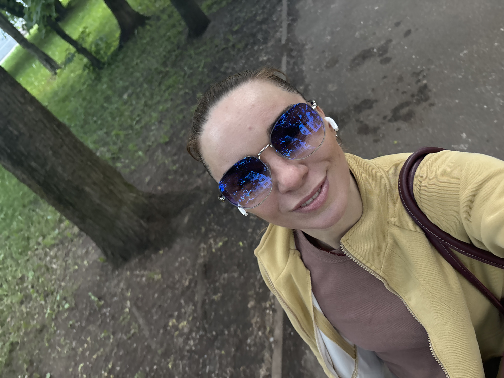 The image is a close-up, high-angle selfie of a woman wearing sunglasses and a yellow jacket. She has a brown top underneath the jacket and is wearing white earbuds. She is outdoors, with trees and grass visible in the background, suggesting a park or natural area. The ground is paved or dirt. The woman is smiling at the camera. The scene appears to be in the city of Sinyavino, Russia.