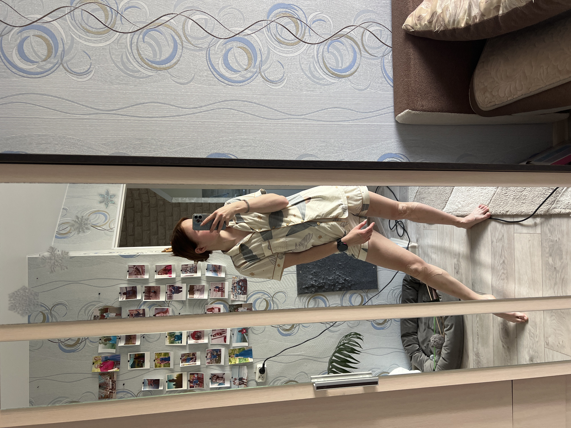 The image appears to be a reflection in a mirror, showing a person taking a selfie in a room.
The person is wearing patterned pajamas and appears to be female, holding a smartphone. The reflection shows the person standing on a light-colored rug.
The room in the reflection has wallpaper with a swirling pattern. There is a sofa with pillows visible in the top portion of the image and also a wall with photographs arranged on it. There is also a plant in a pot visible in the lower part of the image.
The location is presumed to be Chaikovsky, Russia based on the user's request.