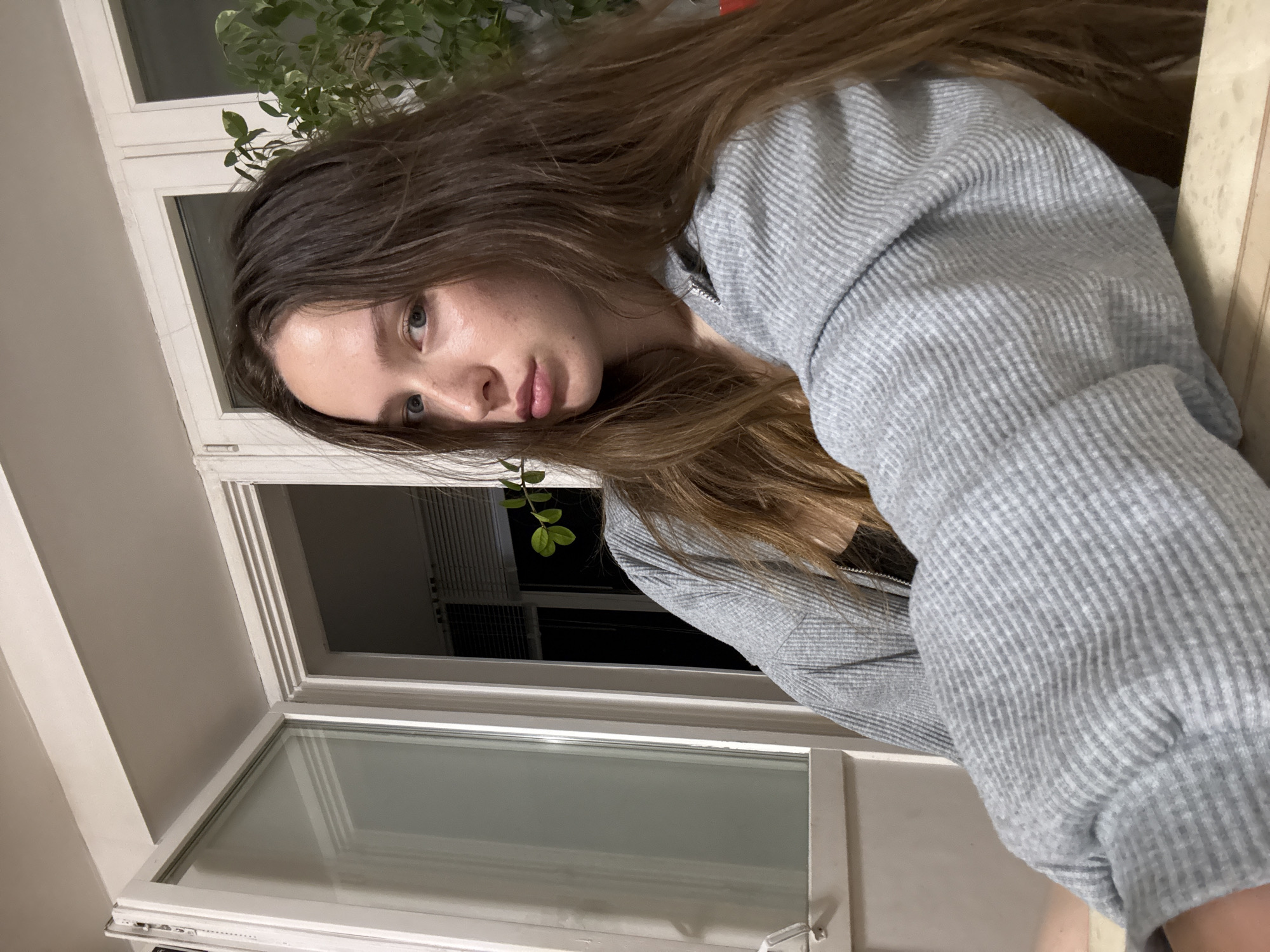 A young woman with long brown hair and blue eyes is wearing a grey, striped sweater. She is positioned in front of a white-framed window with a plant visible behind her, outside the window. The location cannot be determined to be Chaikovsky, Russia from the image alone.
