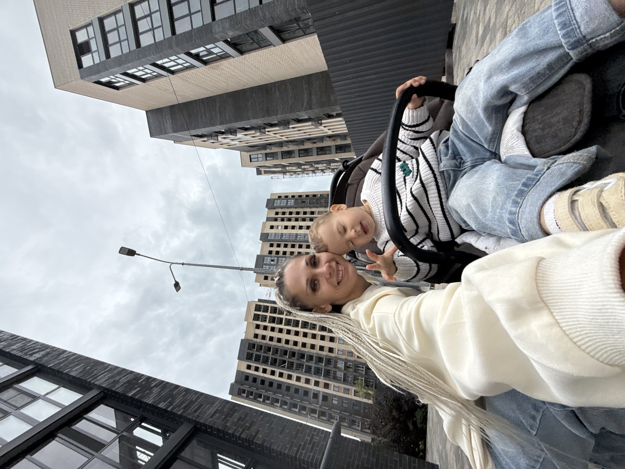 In Rusanovo, Russia, a woman with long, blonde braided hair is taking a selfie with a young child in a stroller. The woman is smiling and wearing a light-colored hooded sweatshirt and jeans. The child, wearing a striped sweater and jeans, is looking down and has their eyes closed. The background features several modern, multi-story apartment buildings under construction and a cloudy sky. The ground is paved.