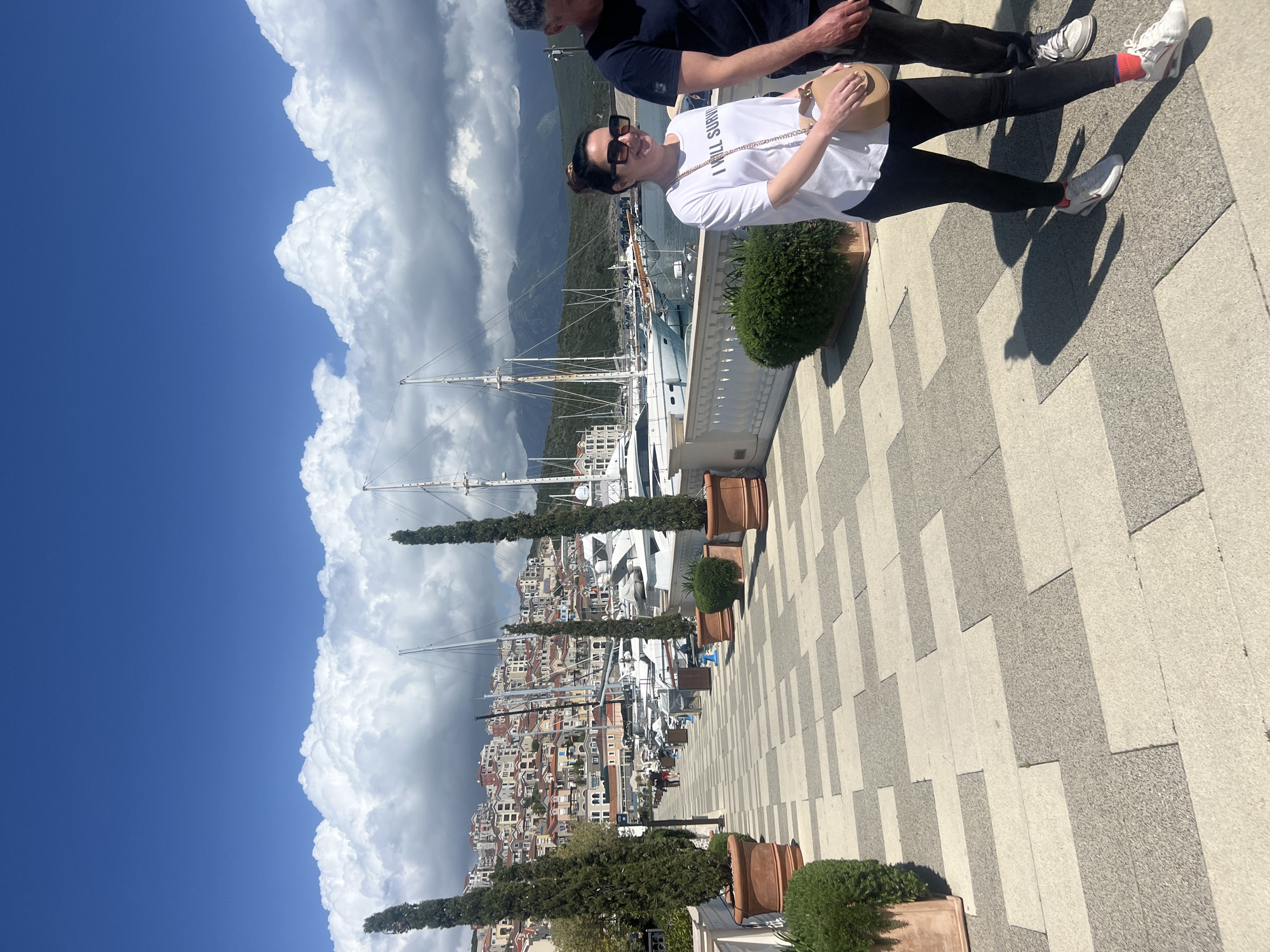 The image shows a woman standing on a checkered sidewalk in Radovići, Montenegro. She is wearing sunglasses, a white t-shirt with a slogan, and black leggings. She is holding a small, light-colored purse. A man is partially visible next to her. The background features a cityscape with boats docked in a harbor, mountains, and a cloudy sky. There are trees in pots along the sidewalk.