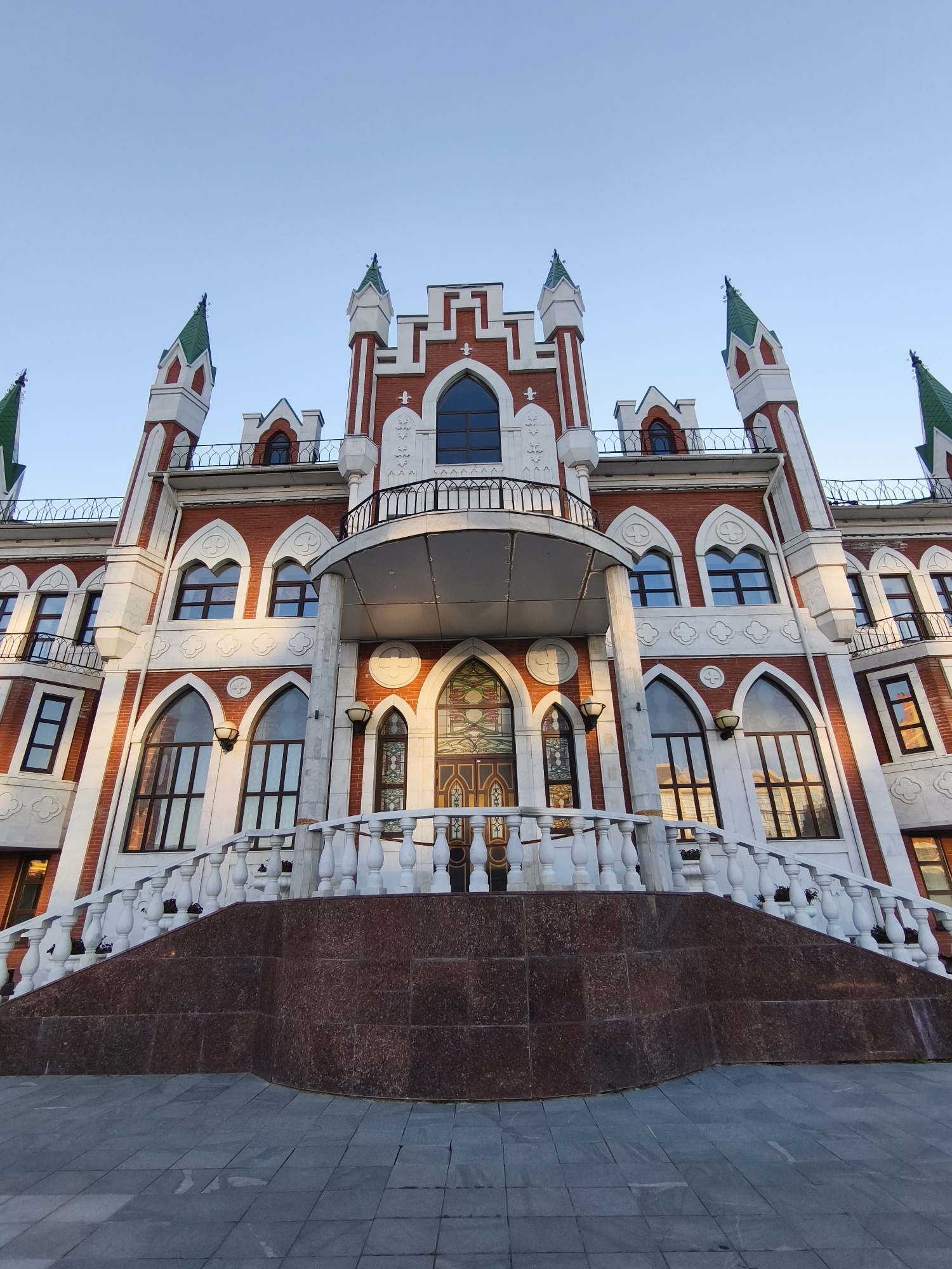 The photograph captures a low-angle view of the Tsar Kokhno Theatre in Yoshkar-Ola, Russia, under a clear blue sky. The building features a red brick facade with white decorative trim and pointed green turrets. A prominent white balustrade with ornate spindles lines a grand staircase leading to the entrance. The staircase is faced with dark red granite. The architecture exhibits gothic revival influences with arched windows and intricate detailing. There are no people visible in the image, and the scene is static.