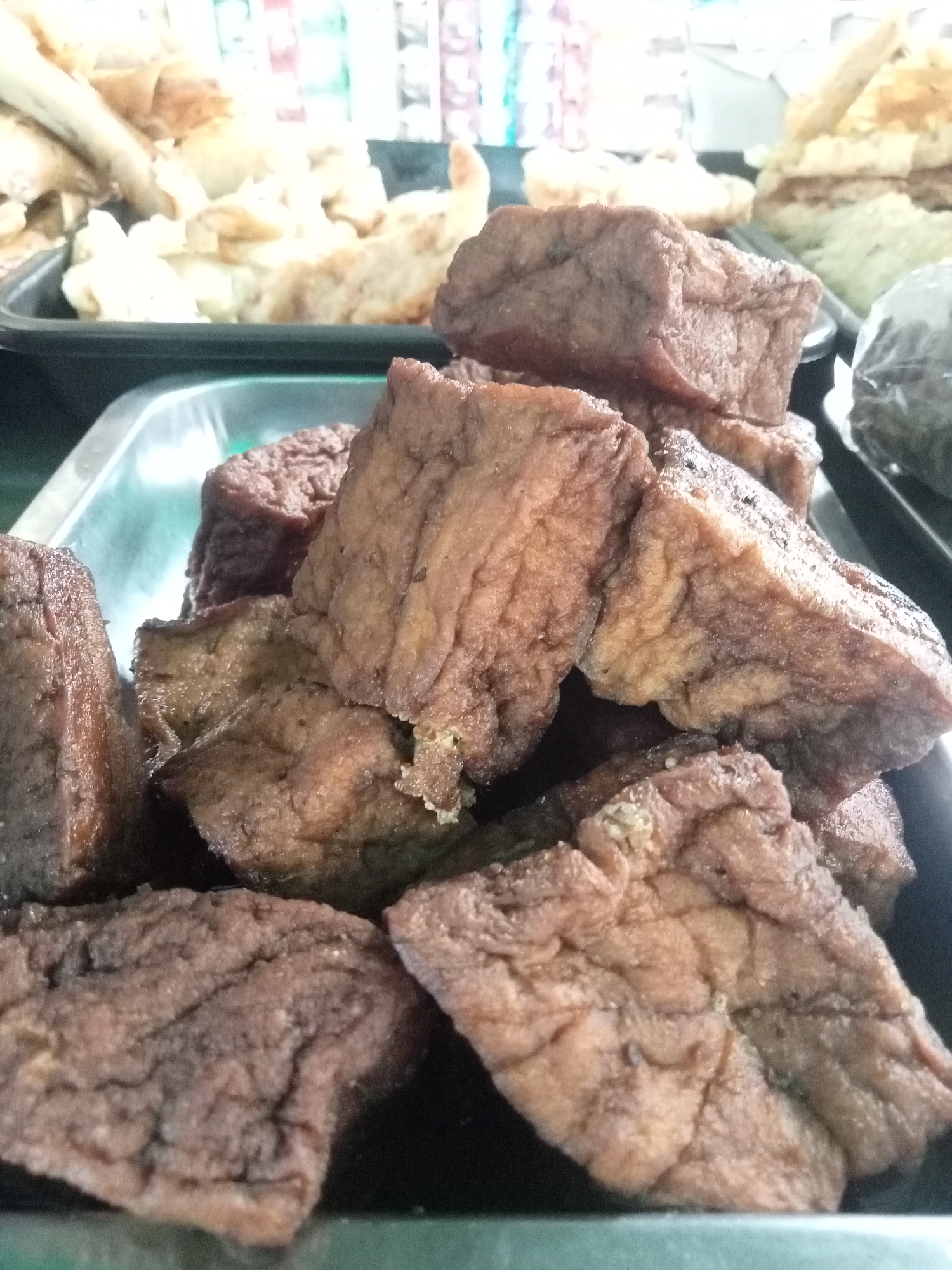 A close-up, overhead shot shows a serving of fried tofu pieces arranged in a metal tray. The tofu pieces are cut into rectangular and square shapes, with a slightly irregular texture. They are golden brown in color, indicating they have been fried. Some pieces appear to be stacked on top of each other, creating a dense arrangement. The background is slightly blurred, but appears to show other food items and possibly packaging, suggesting a market or food stall setting. The lighting is natural and diffused, highlighting the texture and color of the tofu. The image is focused on the food, with no people or other identifiable elements visible. The scene is likely a street food vendor or market stall in Semarang, Indonesia, given the context provided.