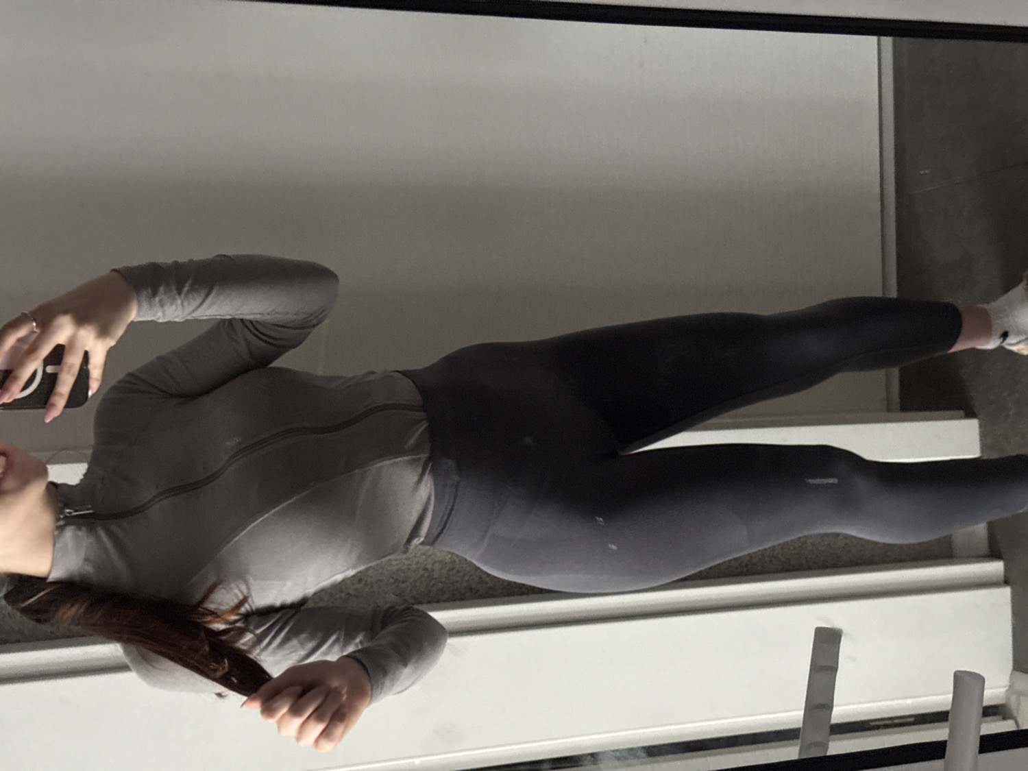 This image, taken in Sofia, Bulgaria, depicts a young woman capturing a mirror selfie indoors. She is shown from approximately the chest to her feet, with a portion of her smiling face and long, dark brown hair visible. She is dressed in athletic wear, including a form-fitting, long-sleeved grey top with a zipper down the front. The top features subtle textured panels. Complementing her top are dark grey, high-waisted athletic leggings, white socks, and light-colored sneakers.
In her left hand, she holds a black smartphone, and a ring is visible on her left ring finger. Her right hand is casually holding a section of her hair. Faint white text, possibly a brand logo, is visible on her left thigh, and the word "GYMSHARK" is discernible on her right thigh. The indoor setting, reflected in the mirror, appears bright with light-colored walls or panels and a darker floor. The woman's slight smile conveys a positive and relaxed demeanor.