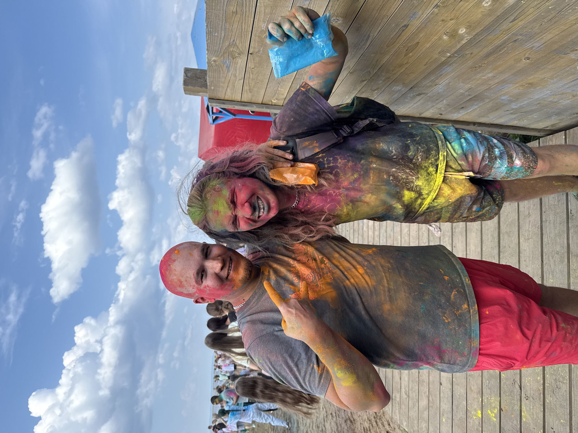 Two young adults, covered in brightly colored powders, stand on a wooden boardwalk. The person on the left, a bald man, is wearing a gray t-shirt and red shorts. He has pink and orange powder on his face and chest, and is pointing his left index finger upwards. The person on the right, with long, wavy hair, is wearing a dark t-shirt and multi-colored shorts. They have green and pink powder on their face and are smiling with their eyes closed. They are holding a small blue packet in their right hand and an orange packet in their left hand. The background shows a beach scene with other people and tents under a cloudy sky.