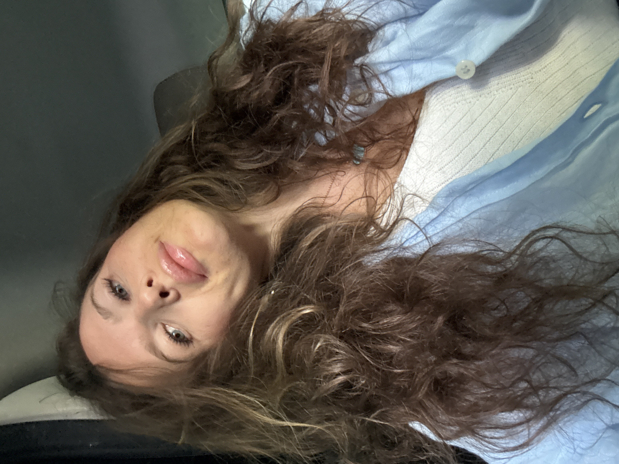 The image captures a close-up portrait of a young woman with long, voluminous, wavy brown hair, seemingly lying down or reclining in an indoor setting. Her head is angled, and she looks directly at the viewer with light-colored eyes (blue or green) and a calm, subtle smile on her full, glossy lips.
She is dressed in a light blue collared shirt, which is open at the neck, revealing a white ribbed top underneath. A small, white button is visible on the blue shirt. A delicate, gold-colored chain necklace rests on her chest, partially obscured by her hair. Her multi-toned brown hair, featuring some lighter highlights, is spread out around her head.
The background is dark and out of focus, suggesting ambient or low-light conditions, keeping the focus primarily on the woman. The lighting on her face is soft and even. No text is visible in the image. The overall impression is one of relaxed intimacy, capturing a casual moment, potentially in Moscow, Russia.