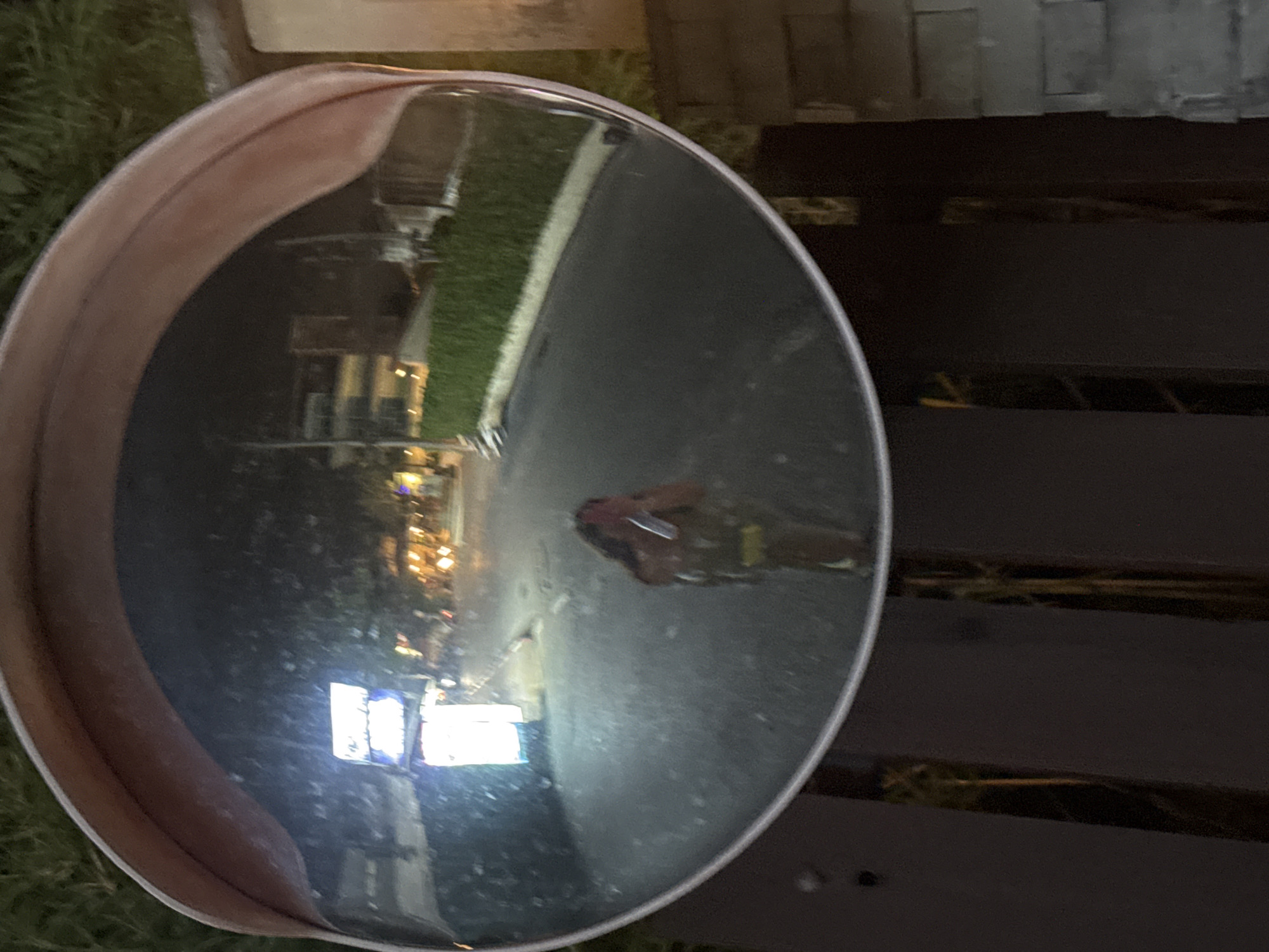 A convex mirror is positioned on or near a dark, horizontal, slatted structure, possibly a bench. The mirror reflects a street scene: buildings are visible in the distance and streetlights illuminate the street. A figure appears to be reflected in the mirror. The street is identifiable as located in Patong, Thailand, due to the building style and lights.