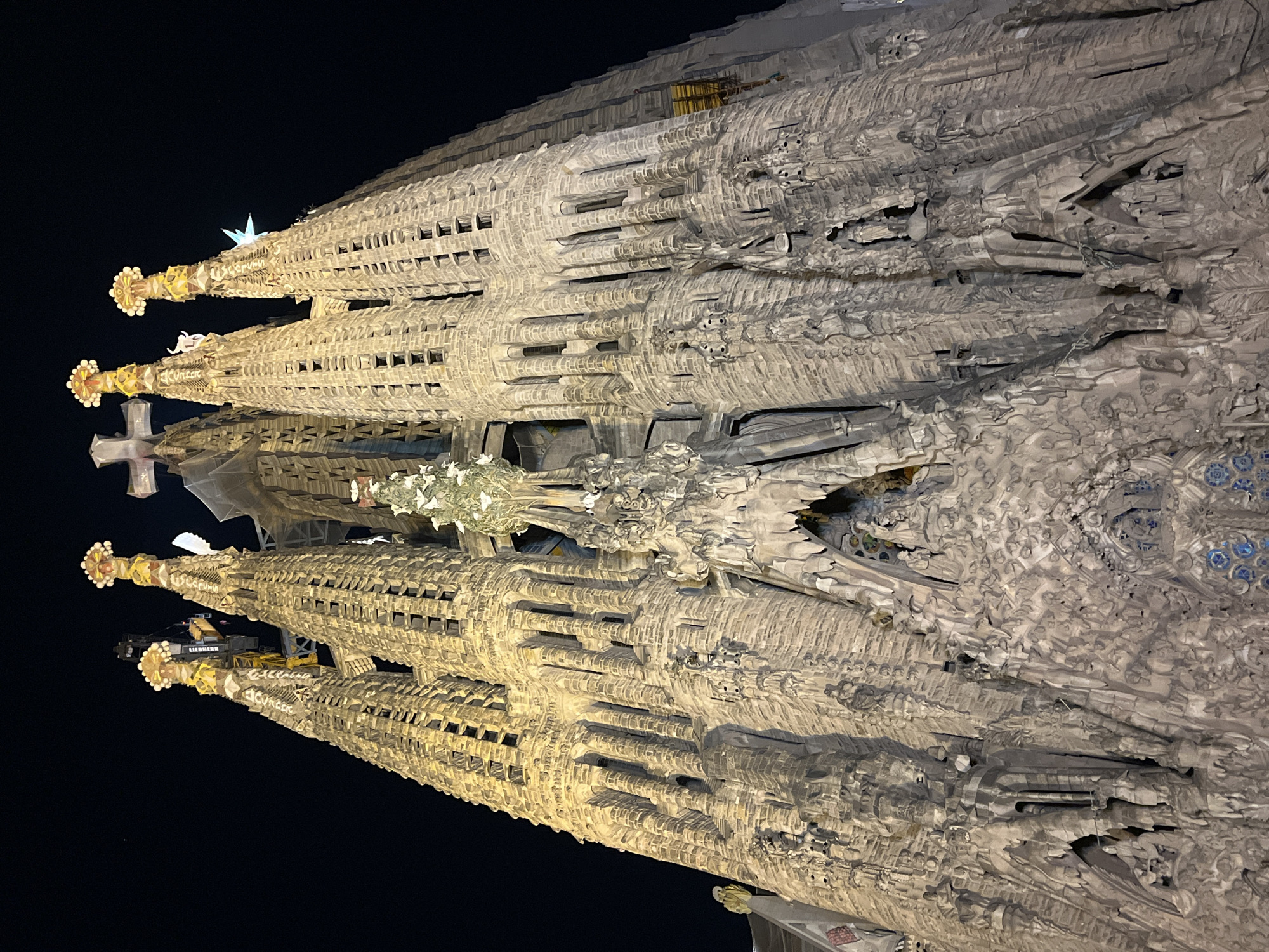 The image captures a detailed view of the Sagrada Familia in Barcelona, Spain, at night. The monumental basilica is illuminated by artificial lights, casting warm, golden tones across its intricate stone facades and towering spires against a dark, black sky.
Multiple tall, slender spires dominate the upper portion of the structure, each tapering to a distinct pinnacle. Several of these spires are adorned with decorative, illuminated elements at their tips, including orb-like clusters in yellow and white, a glowing star-like shape, and a prominent white cross.
The various facades and sections of the basilica are covered in an astonishing array of elaborate carvings and sculptures. These details range from organic, almost melted-looking textures to more defined, relief figures and architectural patterns. The stone surface appears highly textured and complex, with deep shadows and bright highlights emphasizing its three-dimensional quality. Blue mosaic-like patterns are visible in some areas, adding touches of color.
Evidence of ongoing construction is present, with a visible crane, bearing the text "LIEBHERR," and scaffolding integrated into the structure on the right side, highlighting the basilica's unfinished state. Unidentifiable inscriptions are also visible carved into the stone of the spires. The photograph is taken from a low angle, emphasizing the immense scale and architectural grandeur of the Sagrada Familia.