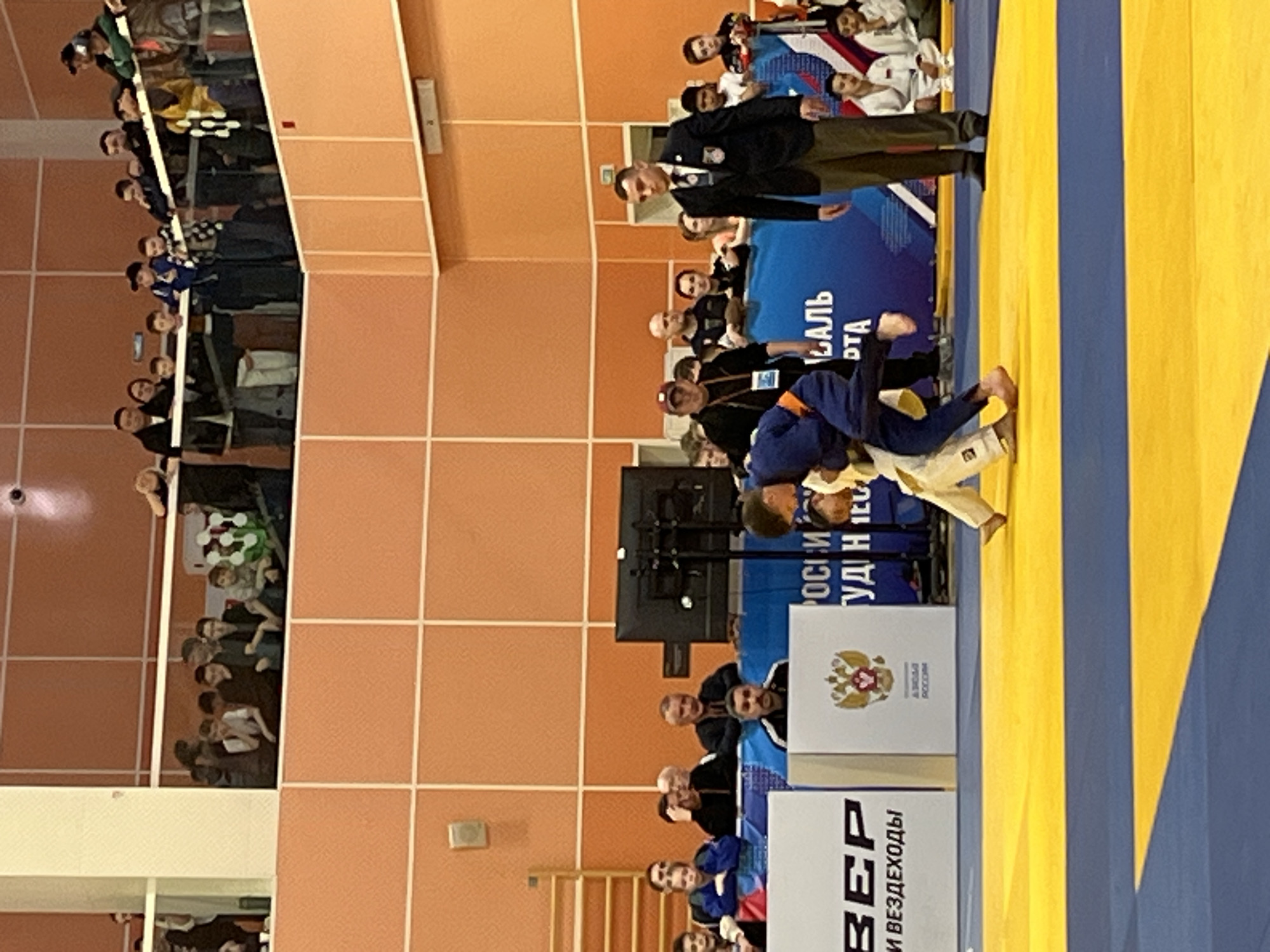 The image depicts a judo match in progress. Two competitors in judogi, one in blue and one in white/beige, are grappling on a yellow and blue mat. A referee in a dark suit and tie is overseeing the match. Spectators are visible in the background, seated in bleachers. The environment appears to be an indoor sports hall with orange tiled walls. Signage in Russian is visible, along with what appears to be the coat of arms of the Russian Federation. The word "Красноярск" (Krasnoyarsk) is visible in the background, indicating that the event is taking place in the city of Krasnoyarsk, Russia.