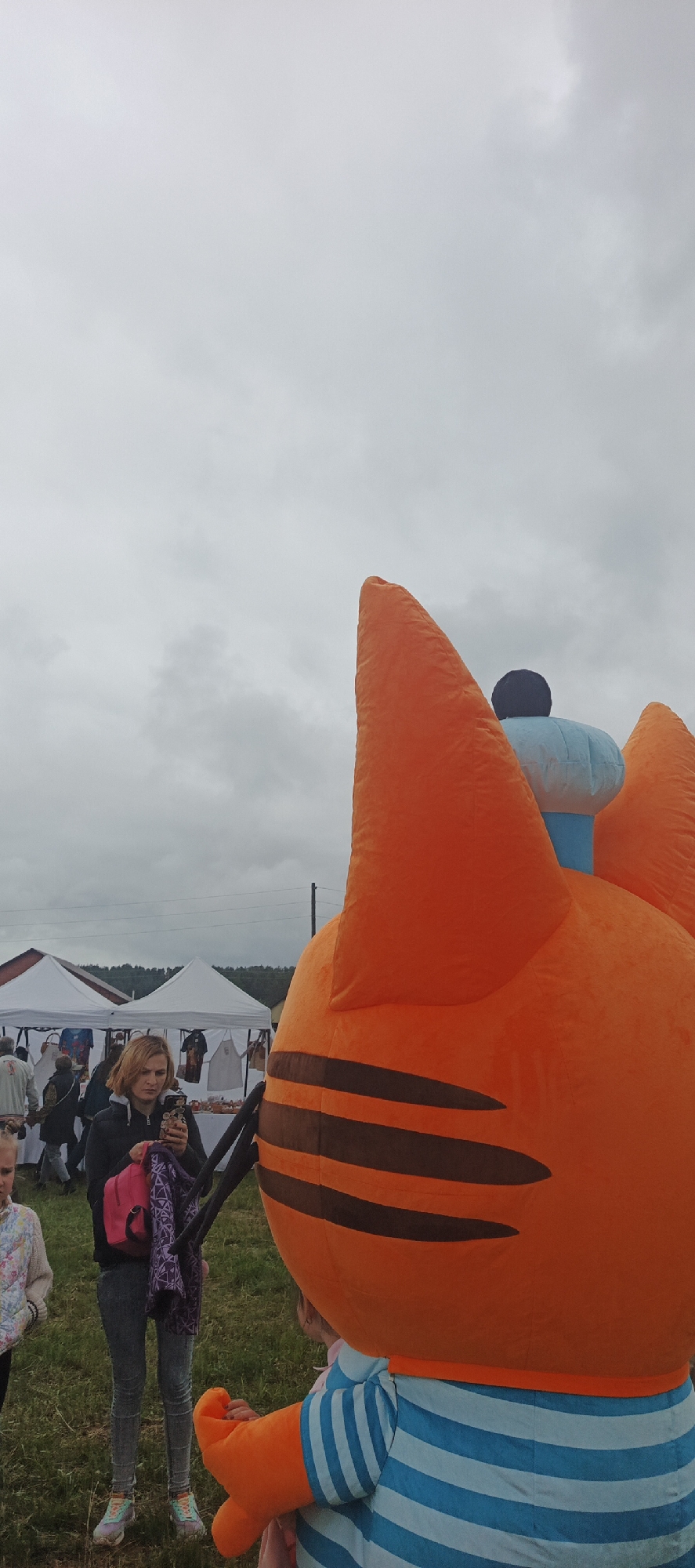 An outdoor scene in Unknown, Unknown, captured under an overcast sky. The foreground on the right features a large, orange inflatable mascot figure, resembling a cartoon cat or tiger. It has dark brown stripes on its orange head and body, with prominent triangular ears. A light blue and black element, possibly a chef's hat, is atop its head. The mascot's torso is clad in a light blue and white horizontally striped shirt. A child's hand is visible touching the mascot's right arm. In the midground on the left, a woman with blonde hair, wearing a dark jacket, light-wash jeans, and multi-colored athletic shoes, stands facing slightly right. She holds a smartphone horizontally with both hands, appearing to take a photograph or video. A pink backpack hangs from her left shoulder, and she grips a purple patterned fabric in her left hand. To the left of the woman, a person with light-colored clothing is partially visible. In the background, several white canopy tents are erected, likely market stalls, displaying various items for sale. Behind the tents, part of a building with a reddish roof and distant trees are visible. Utility lines span horizontally across the upper-left background. The ground is covered in grass.