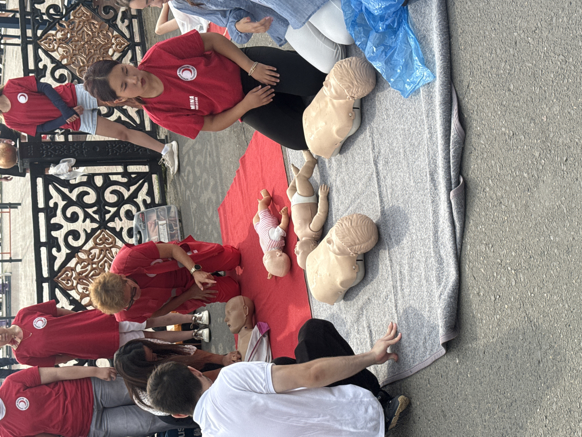 In Pavlodar, Kazakhstan, a group of individuals, mostly wearing red shirts with the Red Crescent Society logo, are participating in a first aid training session. The training involves the use of infant and child mannequins, laid out on a red mat and a gray blanket on asphalt. A man in a white shirt is demonstrating a technique on a mannequin, while an instructor in a red uniform observes and guides. Several other participants, including young women, are kneeling or sitting around the mannequins, observing the demonstration and presumably waiting their turn. In the background, a decorative metal fence and a glimpse of a public area suggest the training is taking place outdoors in a community space.