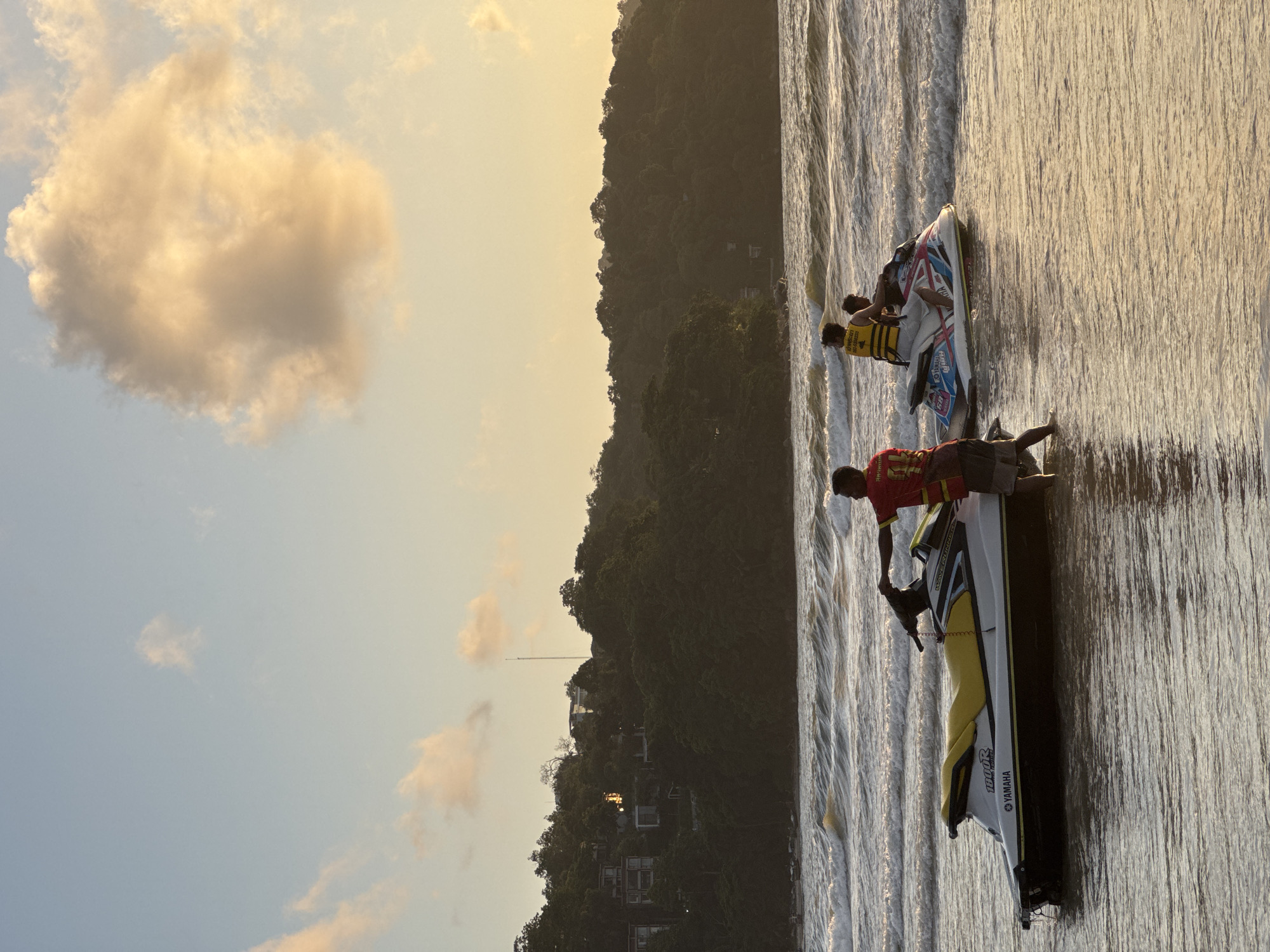 Two jet skis are parked on the shallow edge of a beach, with one person standing beside the front jet ski and another person on the back jet ski. The sky above is a light blue with a few fluffy clouds that appear to be lit by the setting sun. In the background, a lush green hill is covered in trees and scattered with houses. The ocean in front of the beach has moderate waves. The scene suggests a recreational activity at Patong Beach, Thailand, during sunset.