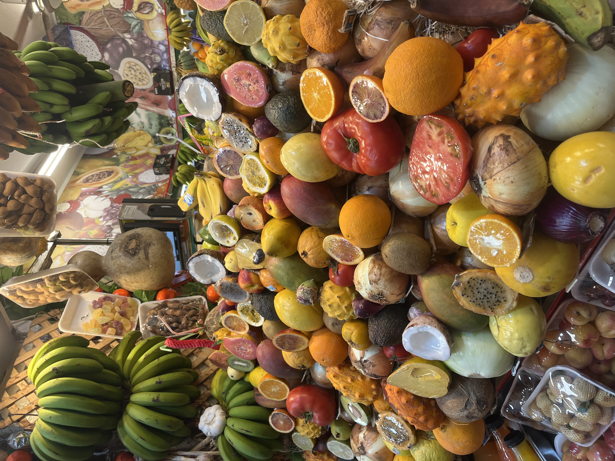 A vibrant and colorful display of fresh produce, including bananas, oranges, tomatoes, kiwis, and other fruits and vegetables, fills the frame at a market. The composition suggests a close-up view of a vendor's stall. The fruits are varied in color, size, and texture, creating a lively visual spectacle. There are packaged foods as well. The market appears to be indoor and there is what may be a menu board and posters above.
The image shows a fruit stand in a market in Las Palmas, Spain.