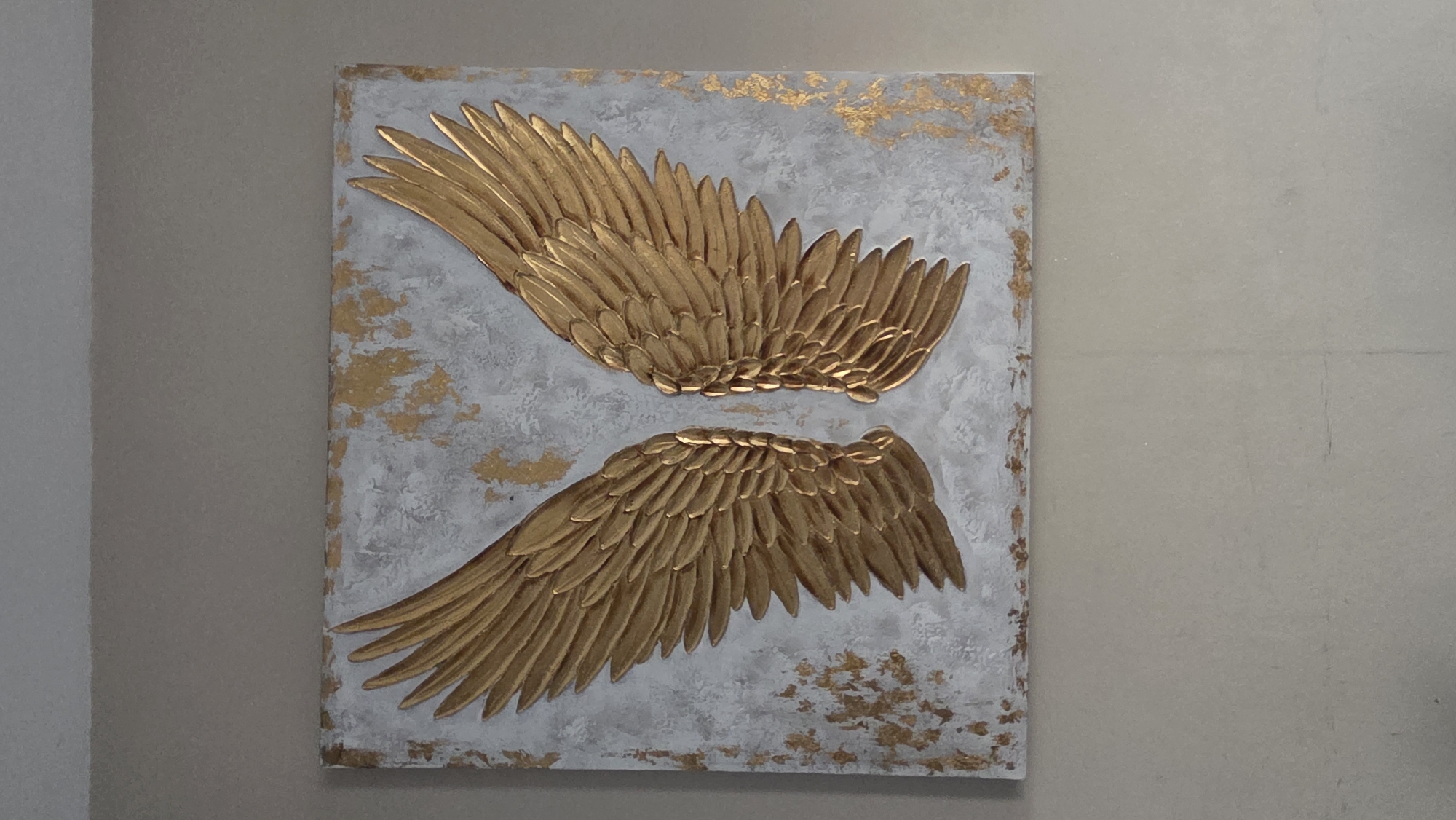 The image shows a square painting of angel wings. The wings are gold and appear to be textured, with individual feathers clearly defined. They are mounted on a white background with flecks of gold leaf scattered across it, creating a distressed or abstract effect. The painting is hung on a plain, neutral-colored wall. The lighting is soft, casting subtle shadows that highlight the relief of the wings. The context of the city of Macé, France is not discernible from the visual information in the image.