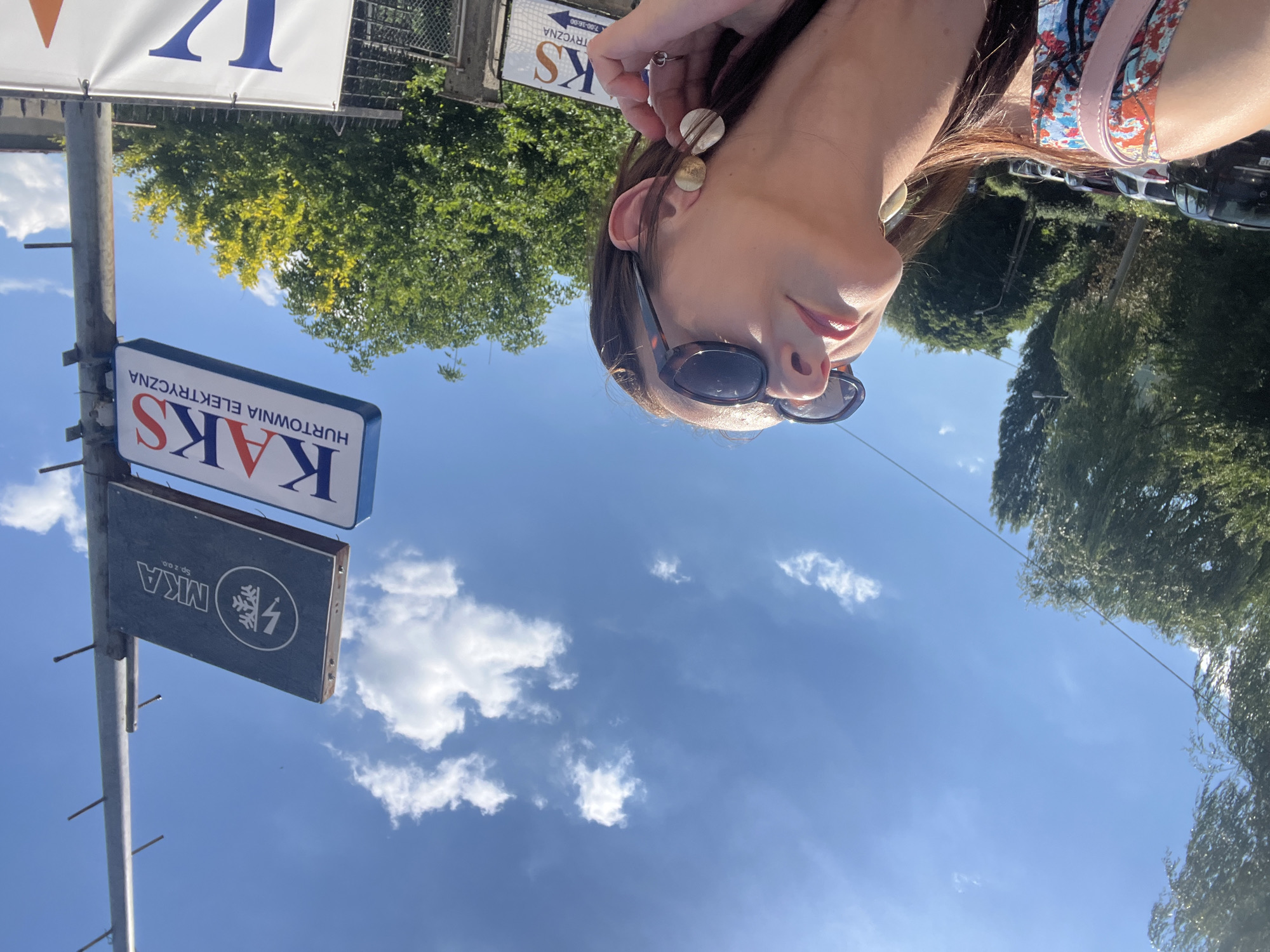 A woman with brown hair and sunglasses is looking up at the sky. She is wearing a colorful top and a pink strap is visible on her shoulder. She is also wearing large earrings and a ring on her right hand. Behind her, there is a sign that reads "MKA Sp. z o.o." and another sign that reads "KAKS HURTOWNIA ELEKTRYCZNA". There are trees and cars in the background, and the sky is blue with some clouds. The scene appears to be outdoors.