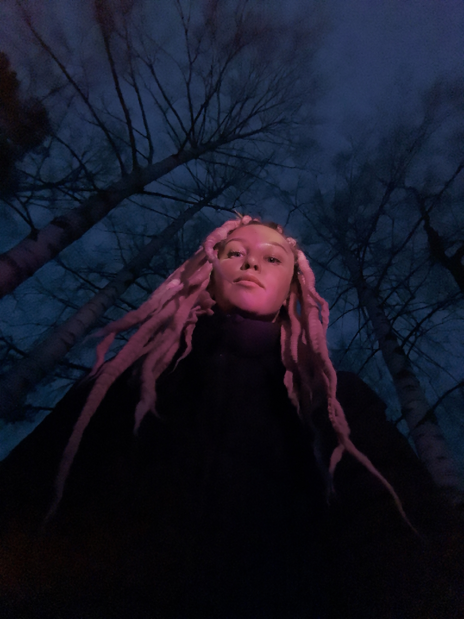 A low-angle, medium shot depicts a person with pink dreadlocks wearing a black turtleneck sweater at night. The person's face is illuminated with a pink light source from below. The background consists of tall, bare trees silhouetted against a dark blue sky. The location is implied to be in Noviy, Russia based on the prompt. The specific environment appears to be a wooded area or park.