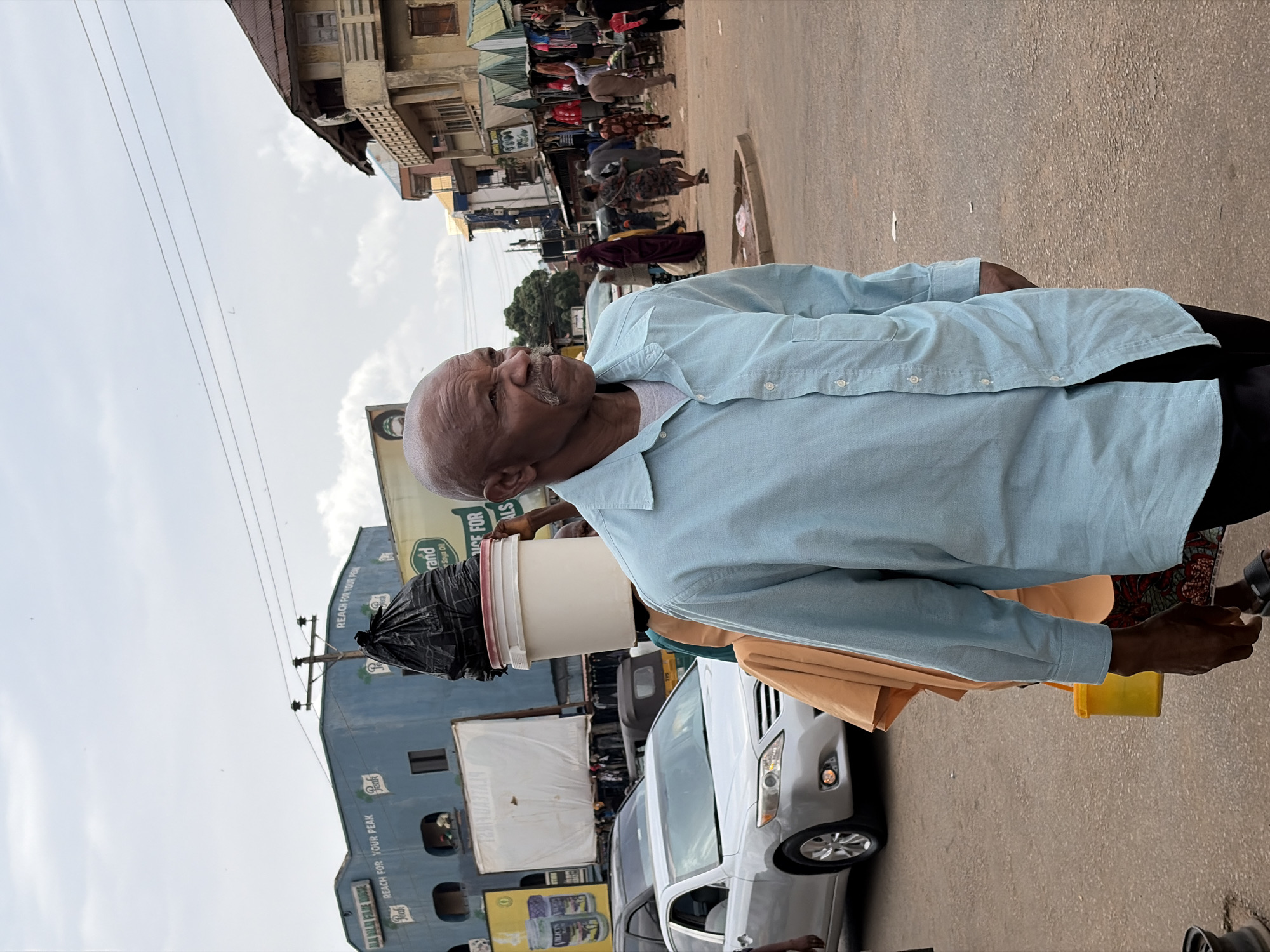 An older Black man with a bald head and a gray mustache walks along a dusty, paved street in Jos, Nigeria. He is wearing a light blue button-down shirt and dark pants. In the background, a woman is walking with a large white bucket and a black trash bag balanced on her head. Several cars, including a silver sedan, are visible on the street, along with various buildings, market stalls, and other pedestrians. The sky is overcast.