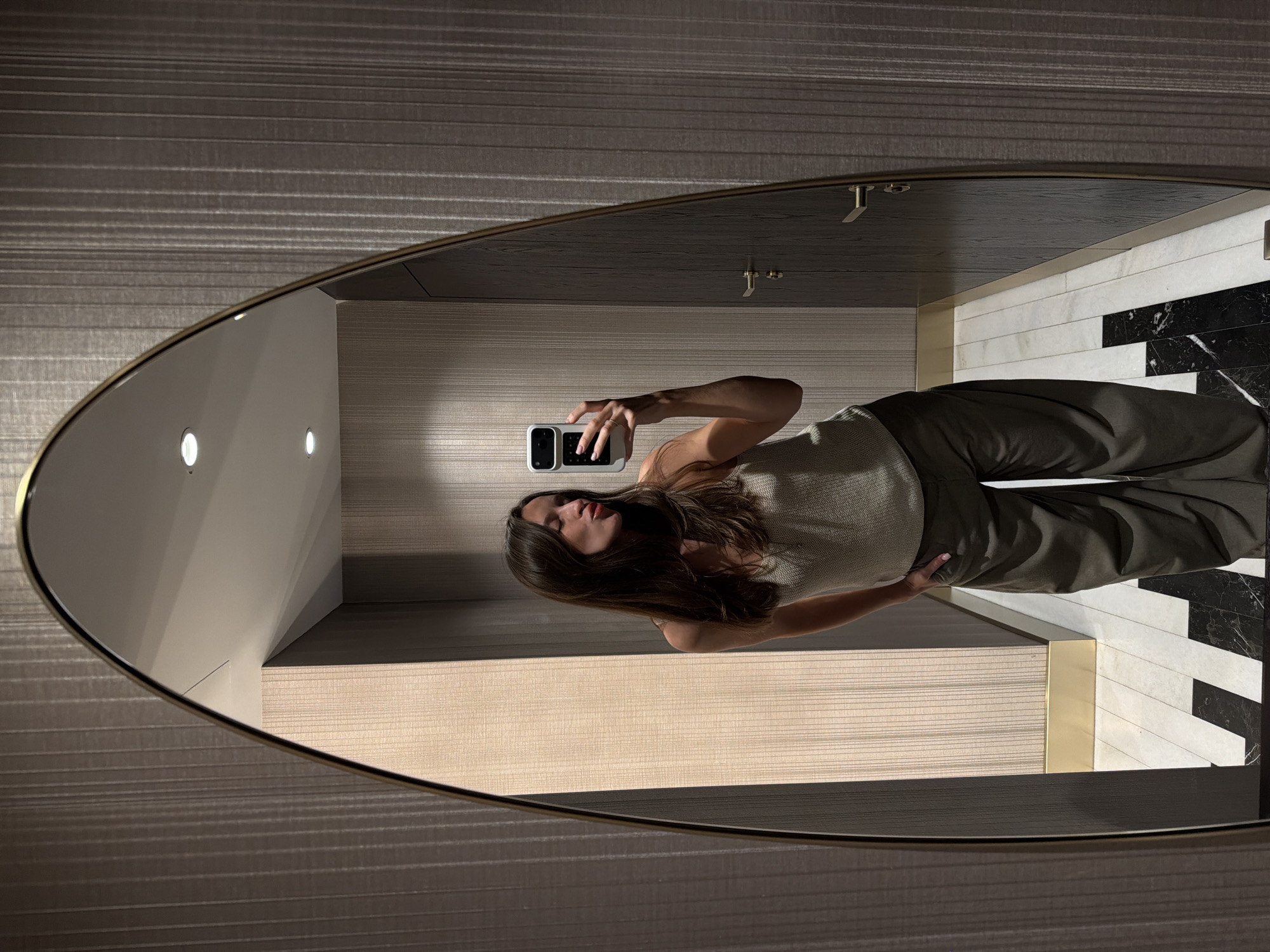 A woman is captured in a mirror selfie within a modern, sophisticated interior space. She is positioned centrally in the oval mirror's reflection, holding a white smartphone with a distinctive multi-button side controller to take the photo. She wears a light-colored sleeveless top and dark, loose-fitting trousers, with her left hand resting on her hip. Her long, dark brown hair falls over her shoulders.
The setting features horizontally striped, textured wallpaper in a neutral shade on the walls. The floor, visible at the bottom of the reflection, showcases a striking geometric pattern of black and white rectangular tiles, some of which have a marble-like appearance. A counter or shelf with brass or gold-colored accents is also reflected. Overhead, several recessed lights illuminate the scene, indicating an indoor environment with artificial lighting. The mirror has a thin, elegant golden or brass-colored frame. There is no visible text in the image.