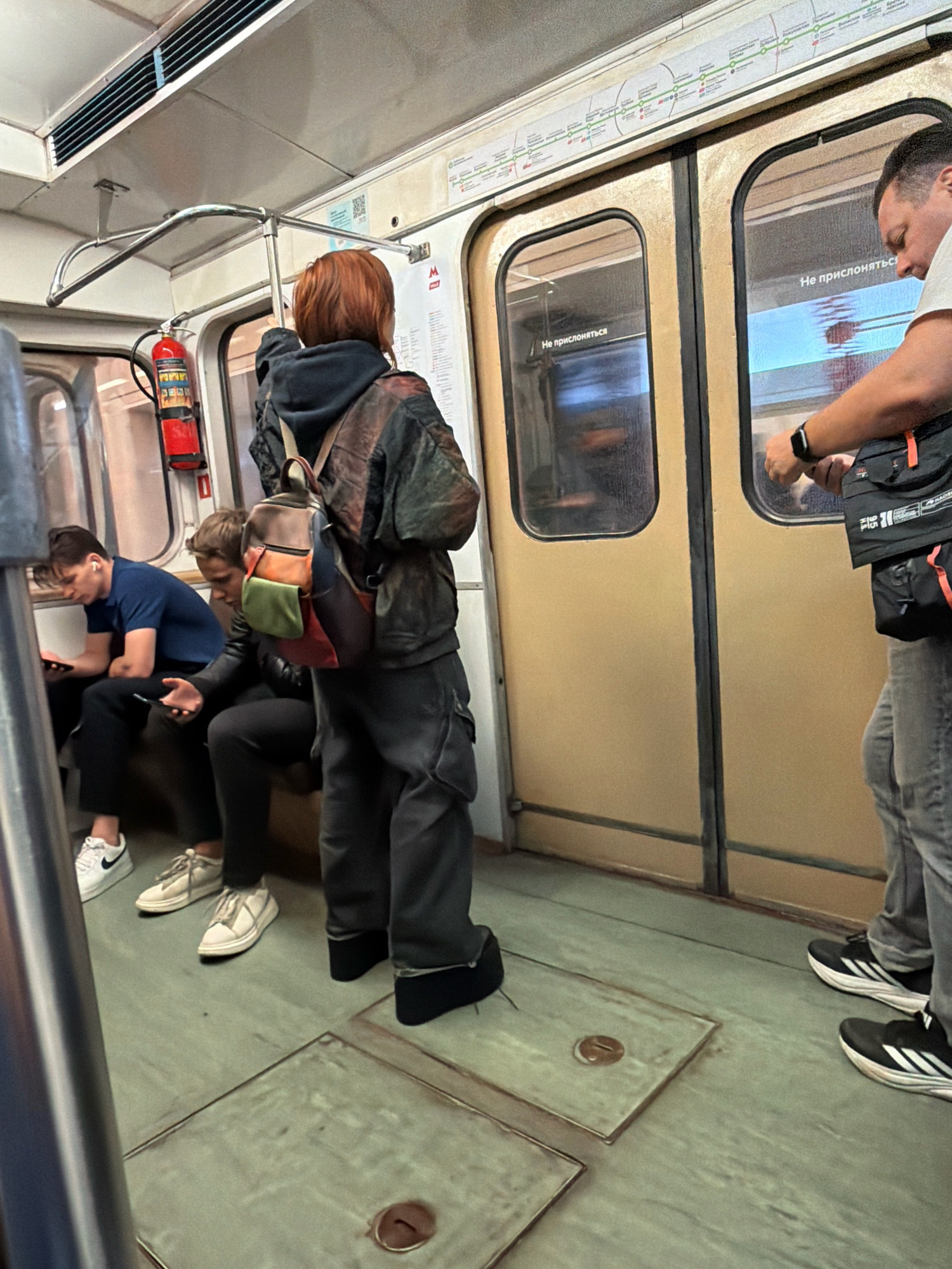 The image depicts the interior of a subway car, likely in Moscow, Russia, given the Cyrillic text visible on the doors which reads "Не прислоняться" (Do not lean). Several individuals are present: a person with short, red hair wearing a dark jacket and a backpack stands facing the doors; a man in a white t-shirt and grey pants, wearing a watch, is standing near the doors; and two men are seated on the bench, both looking at their phones, one wearing a blue t-shirt and the other a dark jacket. A red fire extinguisher is mounted on the wall. The subway car has beige doors with windows and a metal handrail system. The floor is a greenish-grey tiled surface.