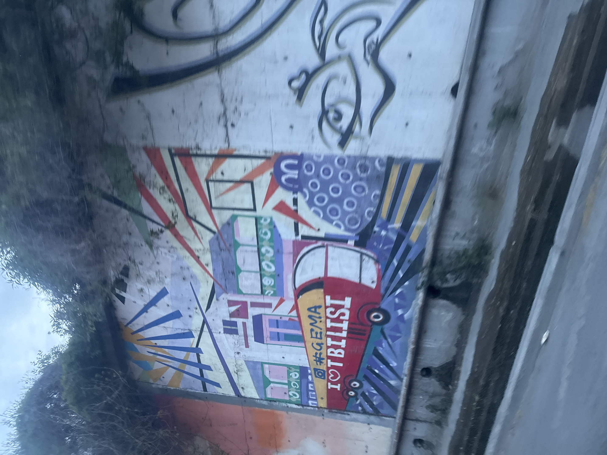 The image shows a concrete wall with a vibrant mural in Tbilisi, Georgia. The mural features a stylized red bus with "I <3 TBILISI" written vertically in white. Above the bus, the hashtag "#GEMA" is displayed. The artwork incorporates geometric patterns in various colors, including blue, purple, yellow, and red. The wall also has other graffiti art, including a black and white abstract drawing above the mural. Green vegetation grows on the left side of the wall. The image is taken from a low angle, looking upwards at the wall. A concrete structure is visible on the right side of the frame.