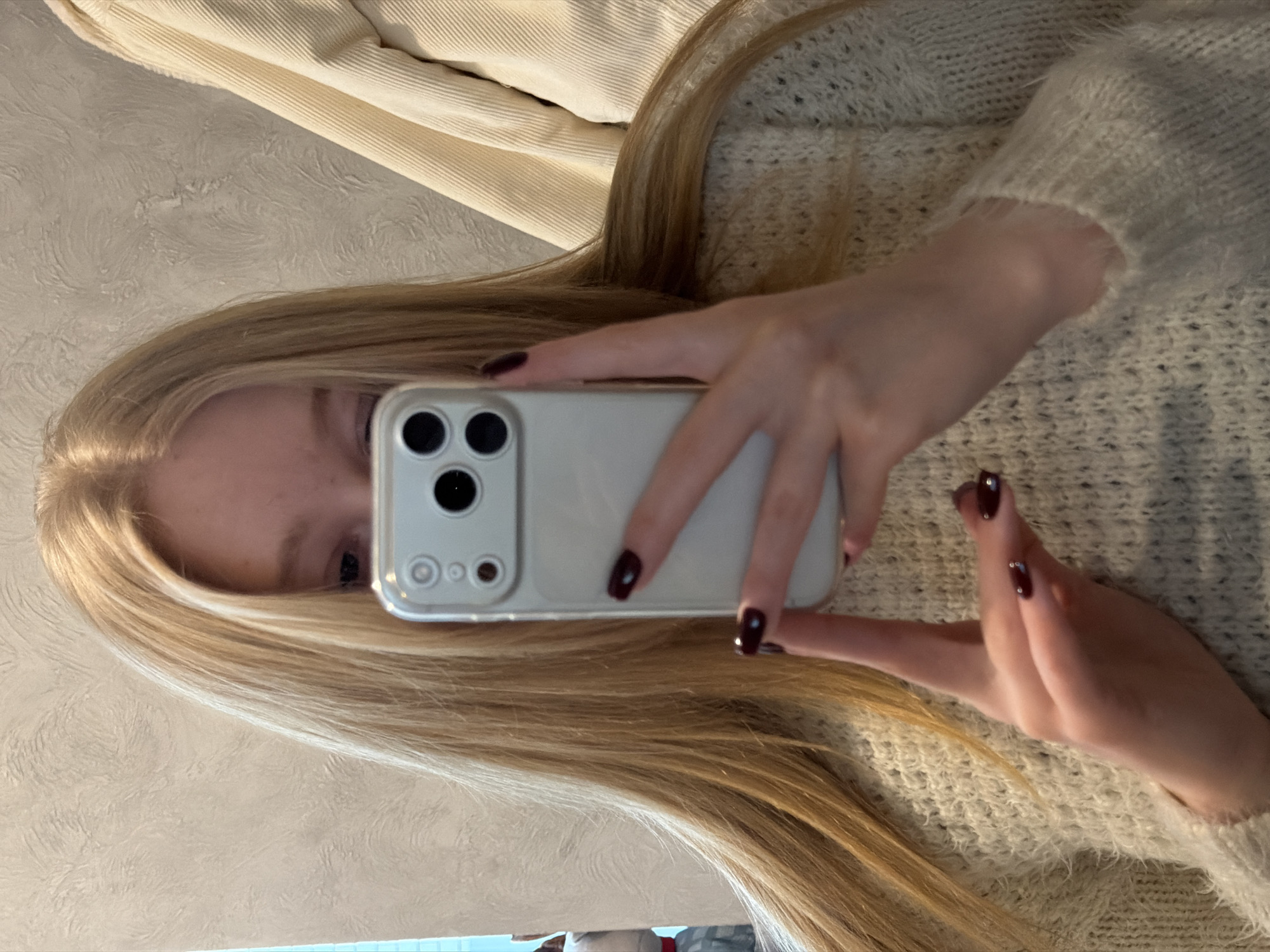 The image depicts an indoor mirror selfie. A person, identified by long, straight, light blonde hair and hands with dark red fingernail polish, holds a light-colored smartphone directly in front of their face, obscuring most facial features except the eyes. The individual is wearing a light beige, textured knit sweater. The smartphone case is light-colored and prominently displays a three-lens camera array with an adjacent flash or sensor. The background behind the person consists of a light-colored wall with an irregular, textured finish. To the right, a segment of a cream-colored, vertically ribbed fabric, likely a garment, is visible. On the far left, a faint reflection indicates a light blue vertical element and possibly another person or patterned object. The specific location within the city of Rossiyskiy, Russia, is not discernible from the visual evidence presented in the image.