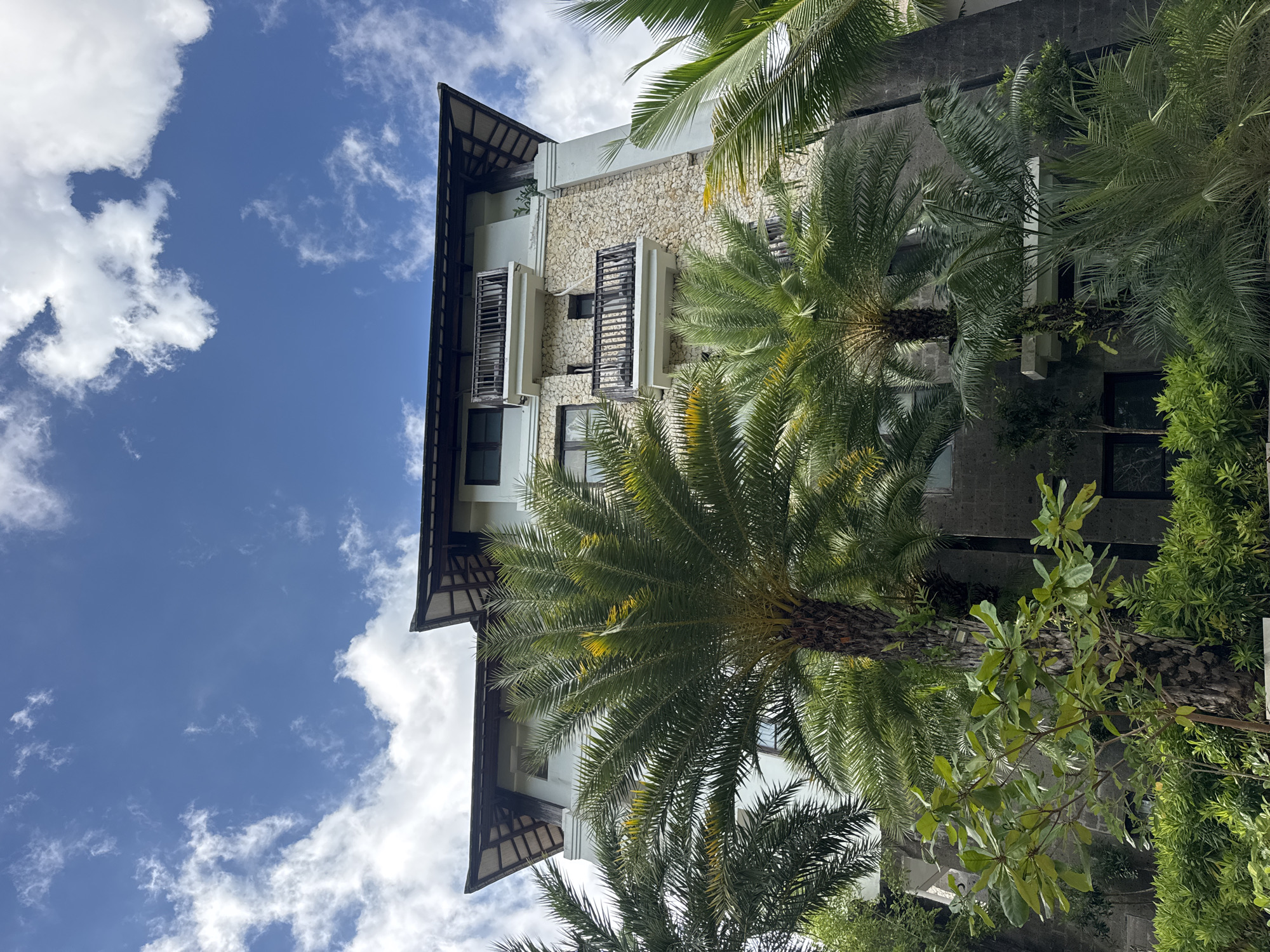 The image shows a multi-story building with a stone facade and a dark, angled roof. Several palm trees are in the foreground, obscuring parts of the building. The sky above is blue with scattered white clouds. The building has multiple balconies with dark railings and windows. The environment appears to be tropical, with lush green vegetation surrounding the structure. The scene is likely outdoors, possibly at a resort or hotel in Kuta Selatan, Indonesia, given the architectural style and vegetation. There are no people visible in the image.