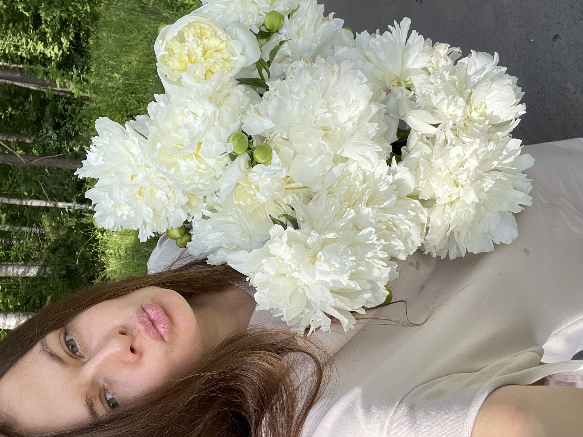 The media file features a close-up, high-angle shot of a woman lying down with a large bouquet of white peonies. The woman has long, brown hair and green eyes, and her lips are slightly parted. She is wearing a light-colored t-shirt. The peonies are lush and full, with some unopened buds visible among the blooms. The background is a mix of green grass and trees, suggesting an outdoor setting, possibly in a park or garden. The lighting indicates it is daytime, likely a sunny day, as evidenced by the bright illumination and shadows. The overall mood appears serene and natural. The photo is taken in Zarechny, Russia. There is no visible text in the media file.