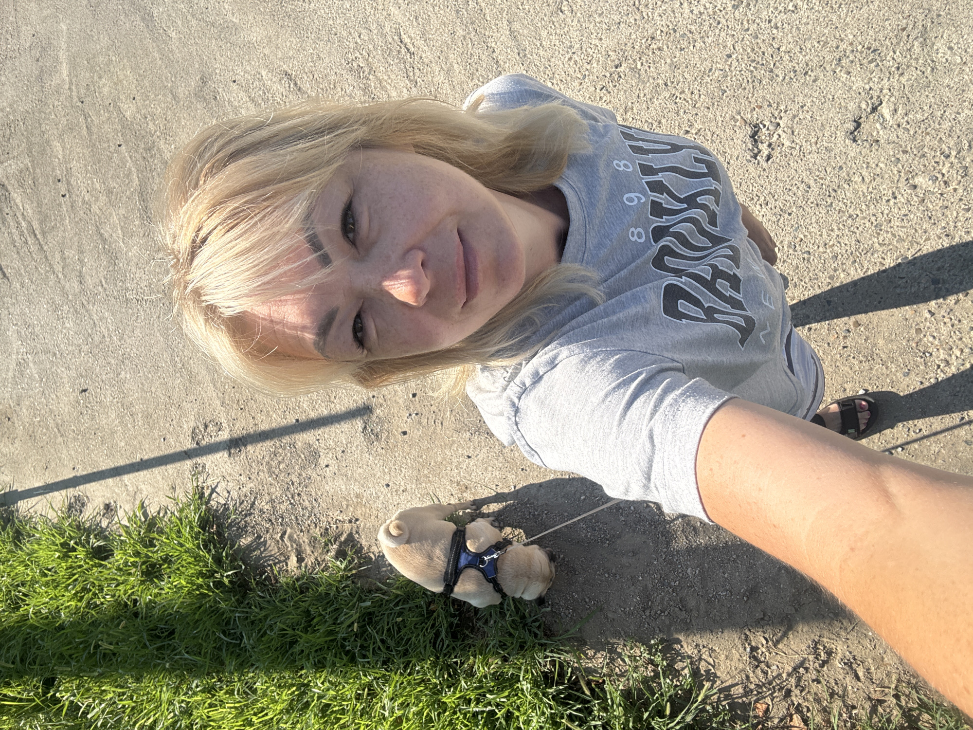 A blonde woman is taking a selfie on a sunny day. She is wearing a grey t-shirt with "BROOKLYN 898" printed on it. She is also wearing grey sweatpants and black sandals. She has a small pug puppy on a leash next to her. The puppy is wearing a blue harness. They are on a sidewalk next to a patch of green grass. The ground is made of gravel and dirt. The image appears to have been taken in a park or along a street in Sochi, Russia. The woman is smiling at the camera, and the puppy is looking down at the ground. The sun is casting shadows on the ground.