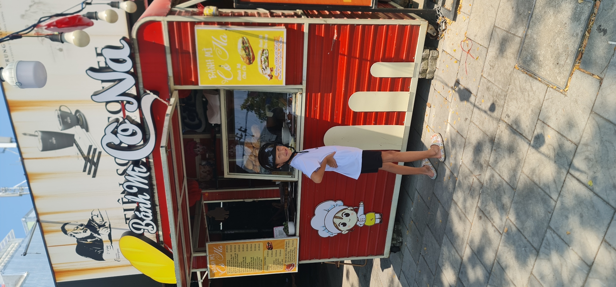 A young boy in a black helmet stands on a paved sidewalk in front of a food cart. The cart, painted with red and white stripes, sells bánh mì. Signs on the cart display images of the sandwiches and list the menu. Text on the cart reads "Bánh Mì Cô Na" and lists a Da Nang address.