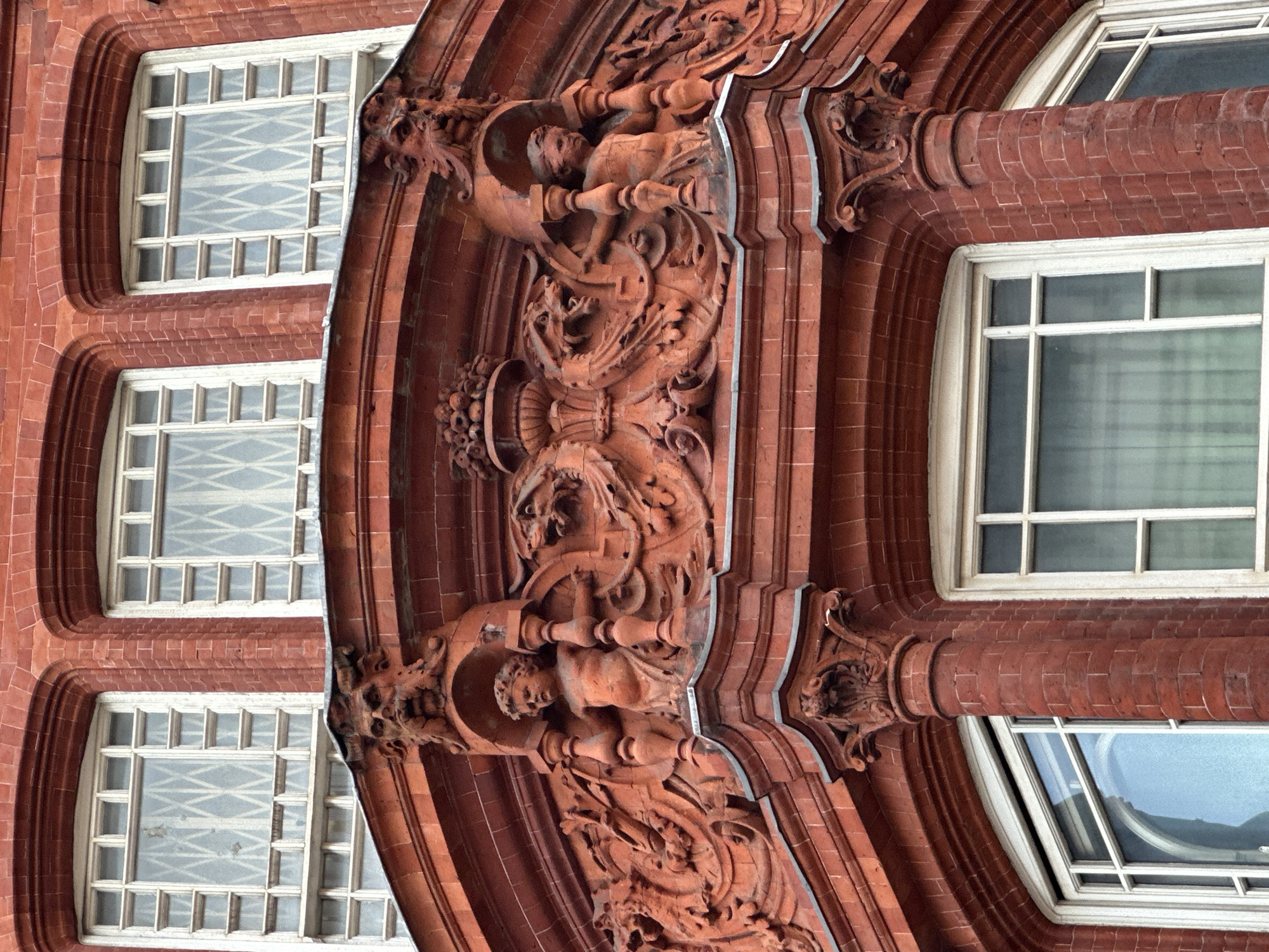 This is a close-up, rotated shot of an ornate red brick and terracotta building, likely from the Victorian or Edwardian era in London. The image is rotated 90 degrees clockwise, showing the building horizontally, but the architectural elements suggest it is an upright structure.
The primary subjects are the building's facade, characterized by its reddish-brown brickwork and highly detailed terracotta ornamentation. Several windows with white frames are visible, many featuring multiple small panes created by muntins. Some window panes reflect a light-colored, crisscross lattice pattern, possibly from interior blinds, while others show reflections of the sky or adjacent structures.
The central focus is a richly carved terracotta column or pilaster. It features an elaborate sculptural relief, including what appears to be a male figure, possibly draped or semi-nude, positioned within an ornamental frame or boat-like structure. Below this figure, intricate scrollwork, foliage, and possibly animalistic forms are carved into the terracotta. The detailing also includes decorative capitals and cornices, exhibiting a high level of craftsmanship typical of the period.
There is no activity or event taking place; the image captures static architectural details. The lighting suggests it is daytime, with soft, possibly overcast light, as there are no harsh shadows. No discernible text is visible on the building's facade. The architectural style, particularly the use of red brick and detailed terracotta, is characteristic of London.