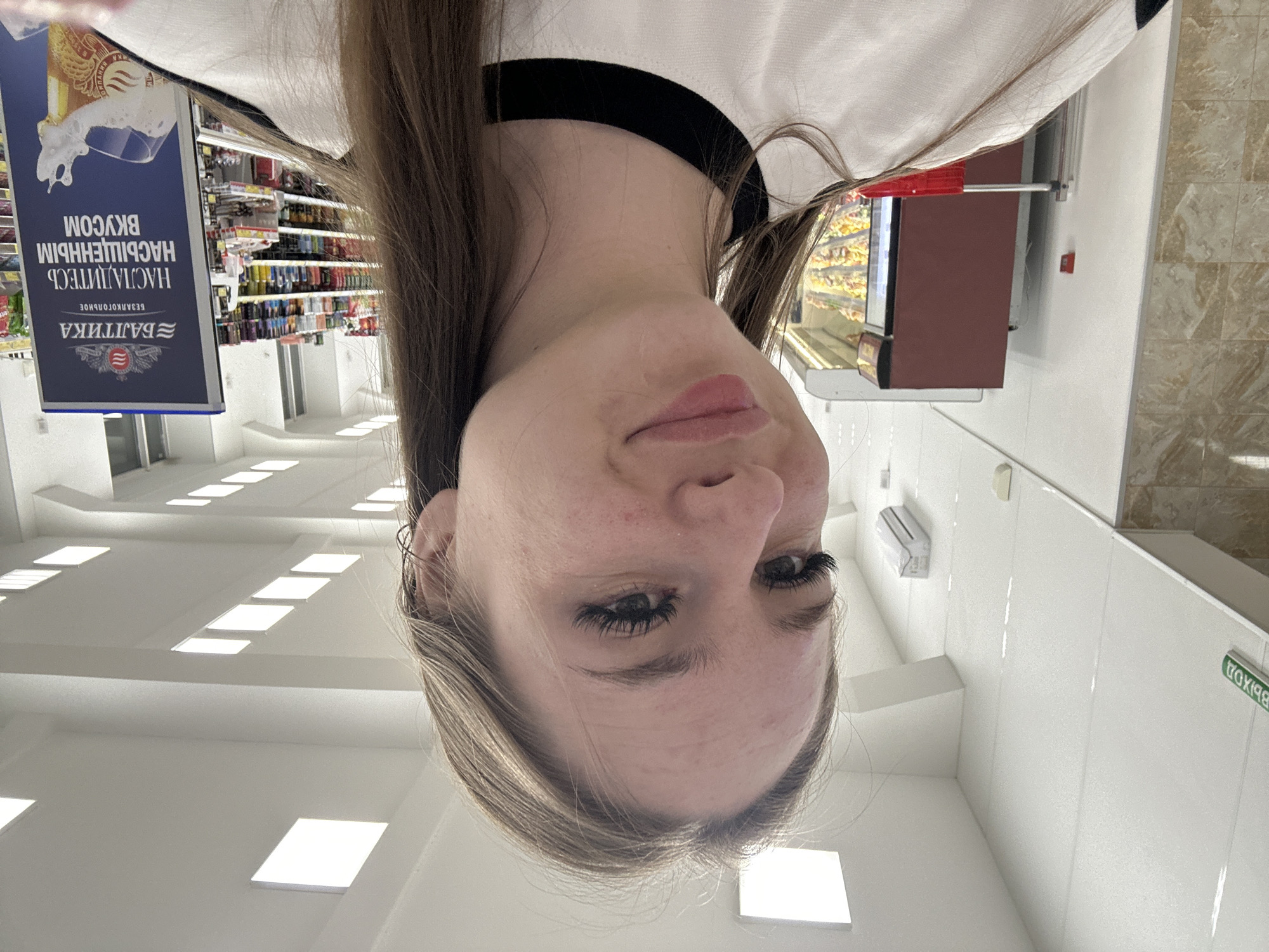 The media file is a slightly overexposed, vertically oriented photograph of a young woman's face and upper body, taken from a low angle. She is looking upwards and slightly to her right, with her head tilted. Her hair is long and brown, and she is wearing makeup, including noticeable eyelashes. The background is out of focus and appears to be an indoor setting, possibly a grocery store or supermarket.
To the left of the woman, a large blue sign with white text is visible. The text appears to be in Cyrillic script and reads "НАСТОЯЩЕЙ ВКУСОМ" (Nastoyashchey vkusom) and "БАЛТИКА" (Baltika). Below this, there are images of a beer glass with foam and a coat of arms.
In the background, shelves stocked with various products, likely beverages, can be seen. To the right of the woman, a display case with food items and a red handle is partially visible, along with a maroon-colored refrigeration unit. The setting is well-lit with fluorescent lighting, suggesting it is daytime, and the interior appears to be a commercial space. The woman's expression is neutral, and the overall mood of the image is casual. There is a green sign with Cyrillic text on the wall to the far right, which is partially obscured but appears to say "ВЫХОД" (Vykhod), meaning "Exit".
