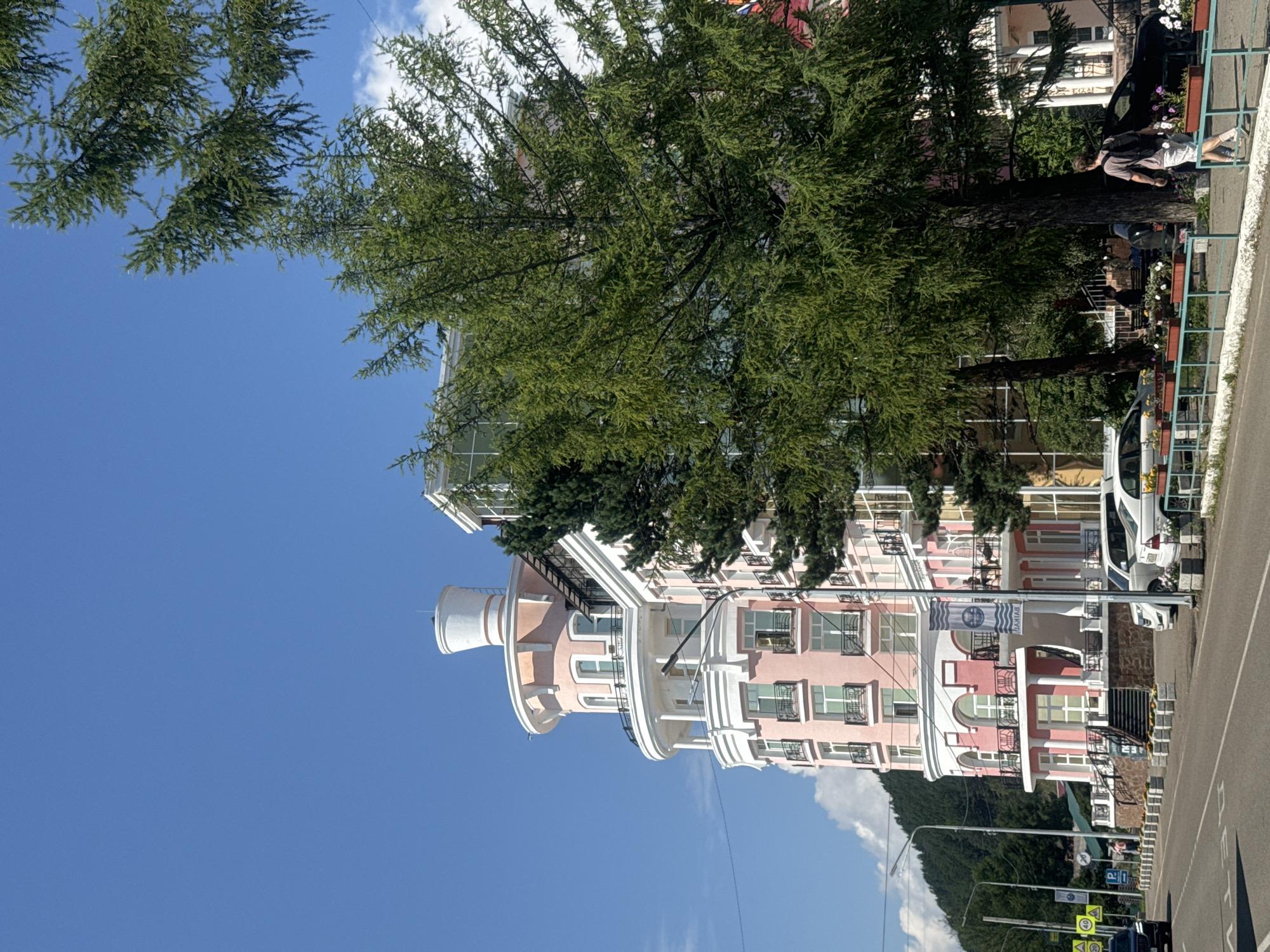 A building with a pink facade and white architectural details is visible on a sunny day in Listvyanka, Russia. The building has a rounded tower section with multiple balconies. A large green tree with dense foliage partially obscures the right side of the building. In the foreground, a paved road runs parallel to the building, with cars parked along the roadside and a pedestrian walking on the sidewalk. The sky is clear and blue with a few wispy clouds. A mountain covered with dark green trees can be seen in the distance.