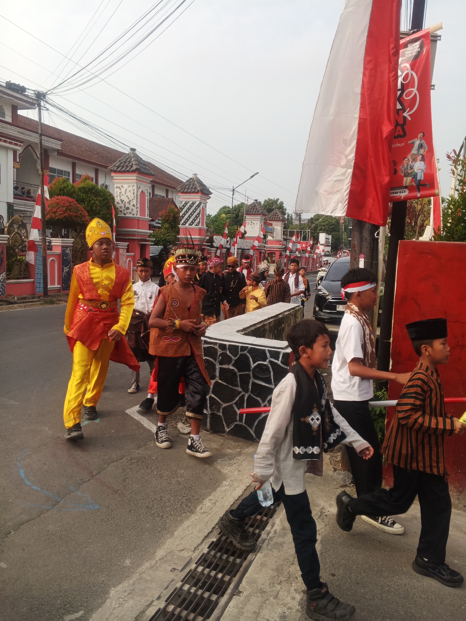 In Sokanegara, Indonesia, a group of young boys in traditional attire are participating in a parade. The boy in the foreground on the left is wearing a bright yellow tunic with orange accents and a matching yellow headdress. To his right, another boy sports a brown vest over a white shirt, with a decorative band across his chest. Behind them, several other children are visible, some in similar traditional outfits with head coverings, while others wear simpler clothing. The parade is taking place on a paved street lined with decorative buildings, featuring red and white architectural elements. Red and white Indonesian flags are prominently displayed along the route, some attached to poles and others hanging from banners. The sky is overcast, suggesting it is either early morning or late afternoon. The overall scene depicts a cultural celebration or a local event.