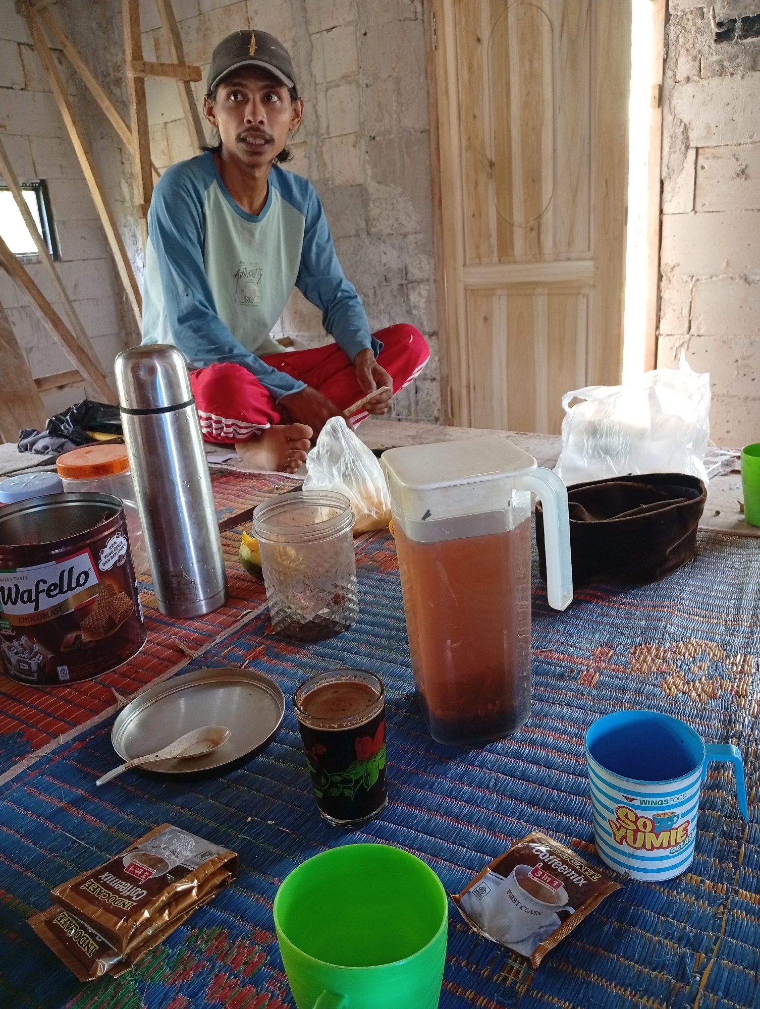 A man wearing a blue long-sleeved shirt, red pants, and a grey baseball cap sits on a blue and red woven mat in what appears to be a construction site. He is looking towards the camera with a slight smile. On the mat in front of him are various items. To his left is a silver thermos. Next to the thermos is a tin of Wafello Chocoblast biscuits. Further to the right, there is a clear plastic jug filled with a reddish-brown liquid, a small glass of dark liquid, and a green cup. A packet of Indocafe Coffeemix is placed near the green cup, and another blue and white striped mug with the words "So Yumies" is also present. Behind the man, a wooden door is visible, and the walls are made of cinder blocks, suggesting an unfinished structure. The overall environment suggests a break time during construction work in Cisitu, Indonesia.