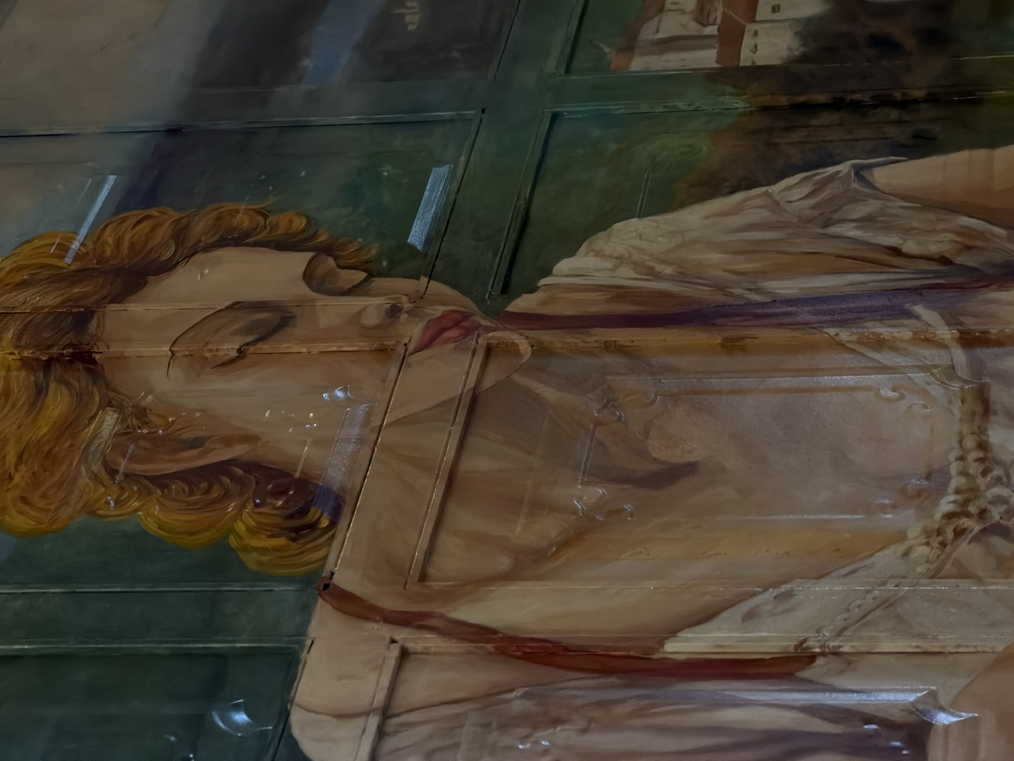 The image shows a section of a painted mural, possibly on a wall or ceiling composed of panels. The mural depicts a reclining figure with fair skin and golden hair. The figure is wearing a draped garment and appears to be sleeping or unconscious. The mural is divided into sections, with a green border between them. The painting style is reminiscent of classical art. The location is Đà Nẵng, Vietnam.