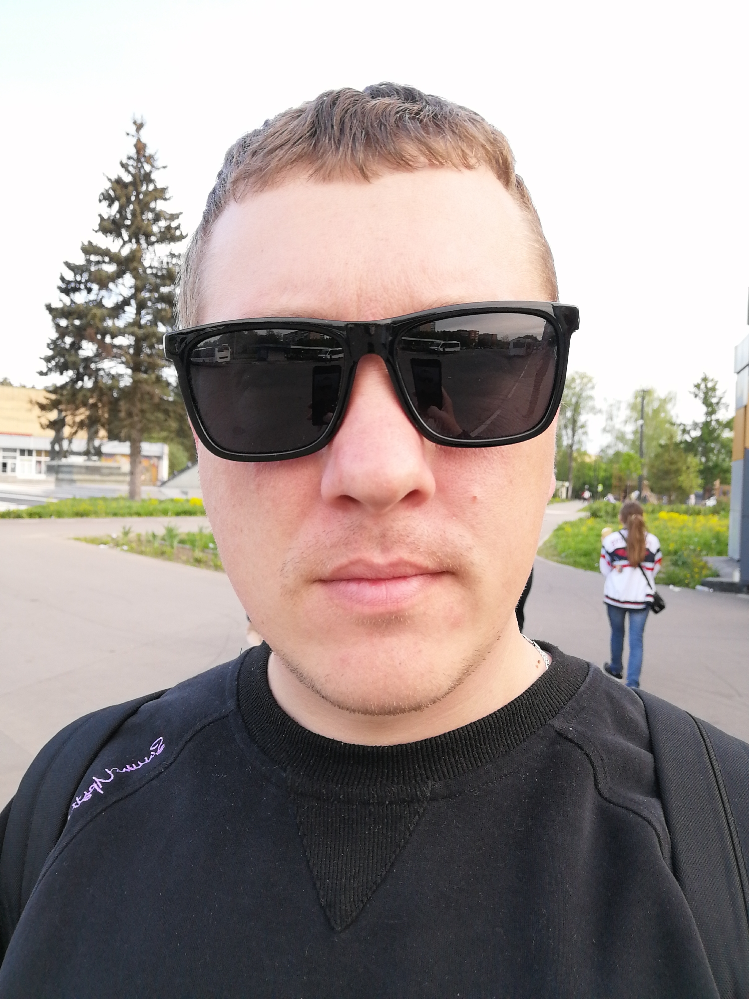 The image is a selfie taken outdoors, likely on a paved walkway. The person taking the selfie is a fair-skinned male with short, light brown hair and stubble. He is wearing black sunglasses and a black sweatshirt with what appears to be purple stitching on the shoulder. He also has a black backpack on.
Behind him, there is a woman walking away. She is wearing jeans and a white jacket with stripes. In the background, there is a tree, a building, and some greenery. The reflection in the sunglasses reveals more buildings and possibly vehicles, indicating an urban setting. The general environment suggests a park or public space in a city. The location is Unknown, Unknown.