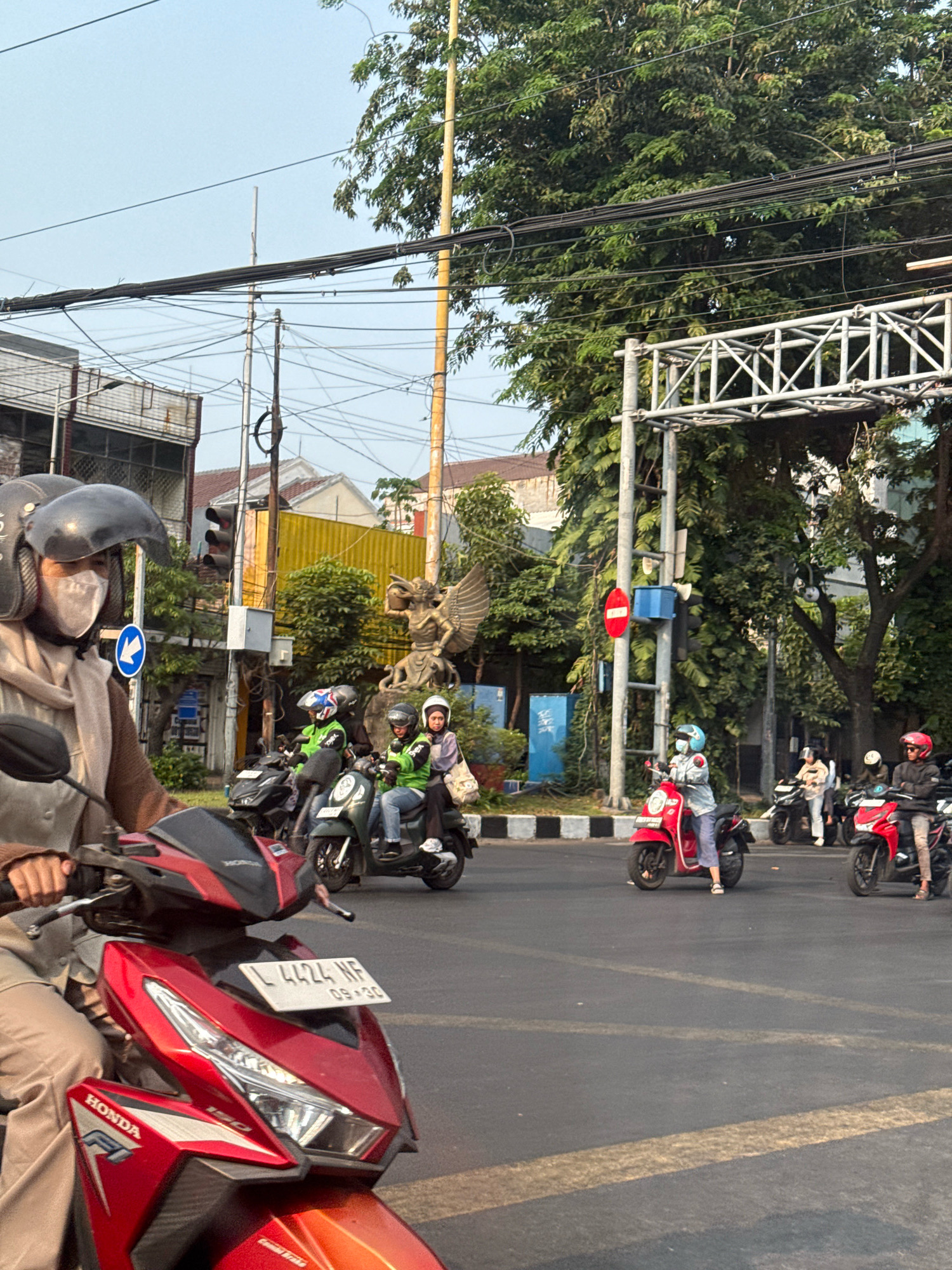 This is a street-level, eye-level, medium shot of a busy intersection in Surabaya, Indonesia. The scene is dominated by numerous motorcycles and their riders, indicative of the city's common mode of transportation. A prominent red Honda motorcycle is in the foreground, with its license plate clearly visible reading "L 4424 WF 09-30".
Several individuals on motorcycles are captured in motion or at a standstill at the intersection. To the left, a woman wearing a helmet, face mask, and a light brown hijab is riding a motorcycle. In the mid-ground, a group of riders are stopped or moving slowly. One rider wears a bright green jacket, another a light purple top, and a third a blue helmet. Further back, more motorcycles and riders are visible, including one on a red scooter and another on a black motorcycle.
The environment suggests an urban setting. Buildings with varied architectural styles, including one with a yellow facade, are present in the background. Overhead, a complex network of electrical wires crisscrosses the sky, typical of a densely populated area. A traffic light, a street sign with a blue arrow, and a red circular traffic sign are visible. A large green tree provides a natural element to the urban landscape, and a stone sculpture, possibly of a mythical creature, stands on the right side of the intersection. The road surface is asphalt, marked with yellow lines. The overall impression is a snapshot of daily life and traffic flow in a bustling Indonesian city.