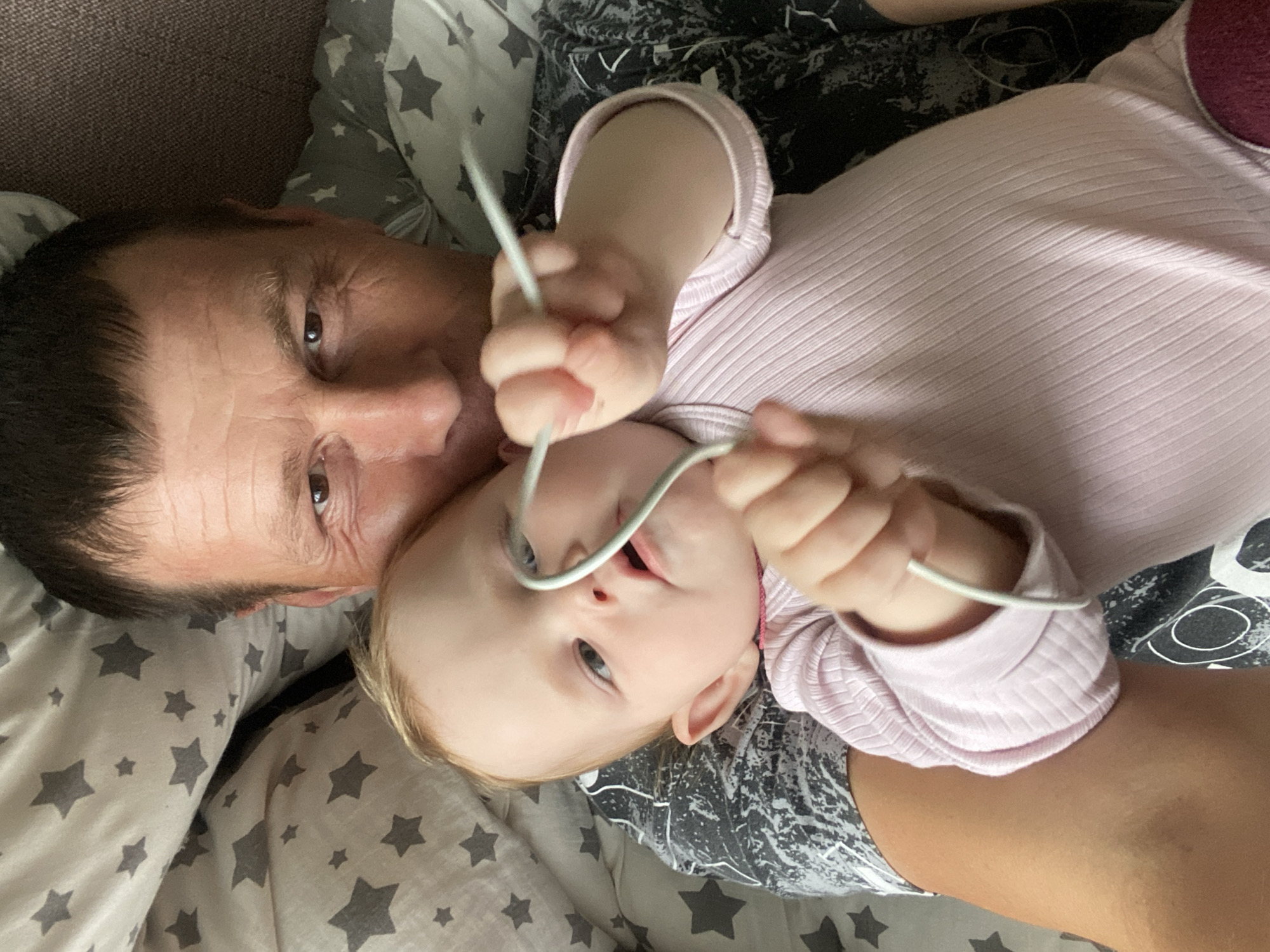 The image captures an intimate, overhead view of an adult male and a baby lying close together, likely on a bed or sofa. The man, positioned on the left, has dark hair, brown eyes, and visible wrinkles on his forehead. He is lying on his back, with his head turned towards the baby.
The baby, appearing to be an infant or toddler, is central to the frame, facing the camera directly. The baby has light blonde or light brown hair and curious blue or grey eyes, and is wearing a light pink, ribbed long-sleeved top. The baby is actively engaged, holding a thin, white or light grey flexible cable with both hands and bringing it towards their mouth, characteristic of a young child exploring objects.
The background consists of soft furnishings: a grey and white star-patterned pillow or sheet is visible, along with a darker, mottled or abstractly patterned fabric beneath the baby. The lighting is soft and even, suggesting an indoor setting. The scene conveys a sense of warmth, family bonding, and the playful curiosity of the baby. There is no visible text, and the indoor setting does not provide cues for weather or a specific time of day. While the location context is Фоки, Russia, this is not discernible from the image itself.